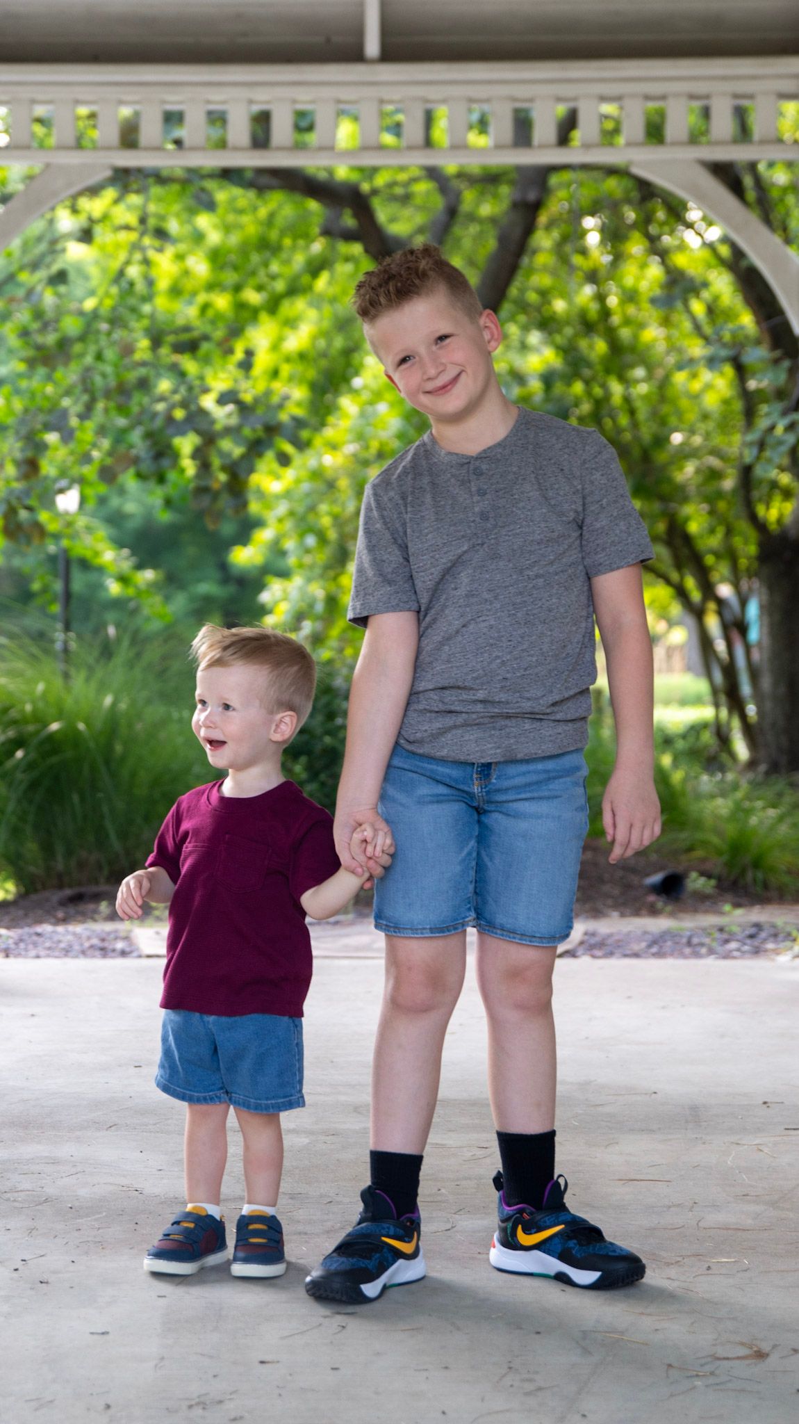 Two children in casual wear stand together on a concrete path surrounded by greenery.