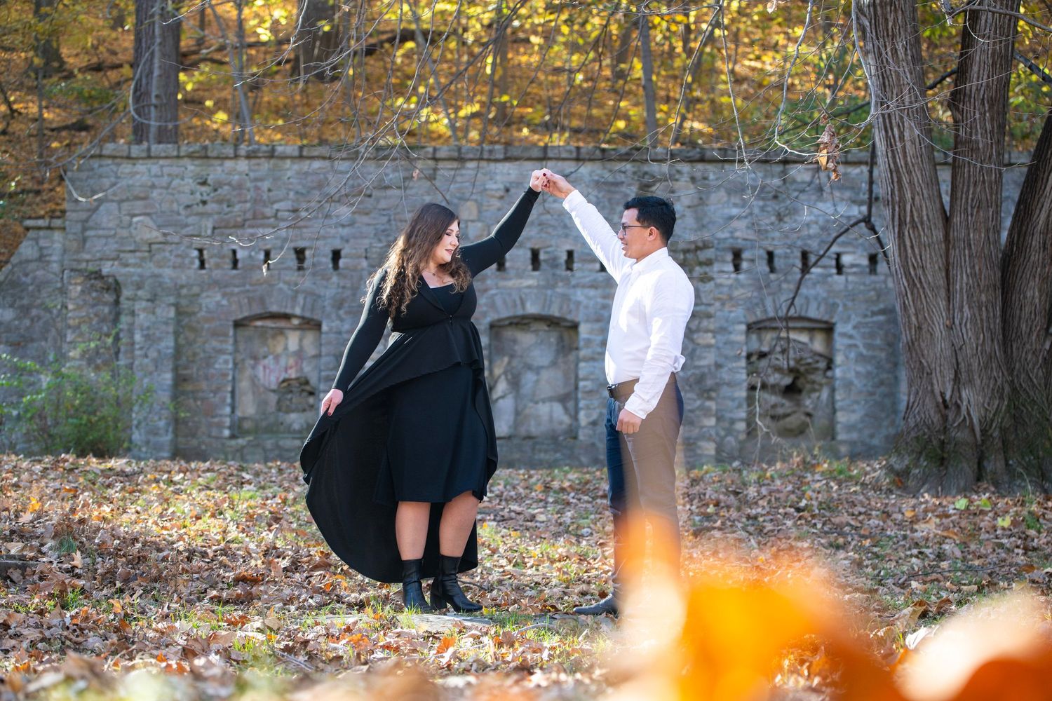 Couple poses playfully against stone wall surrounded by autumn leaves in romantic outdoor photoshoot.
