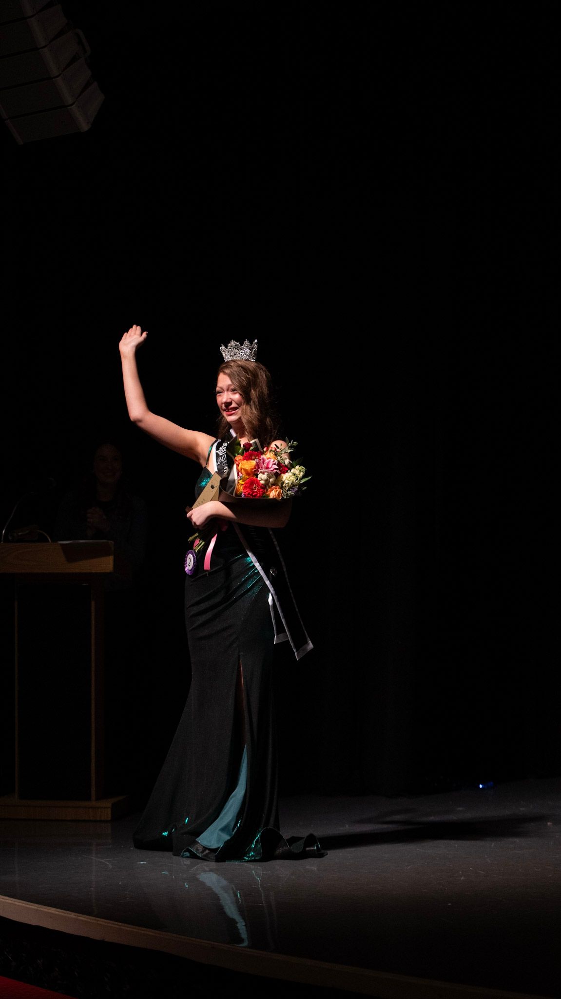 A pageant winner in a black gown celebrates with raised arms on a dimly lit stage.