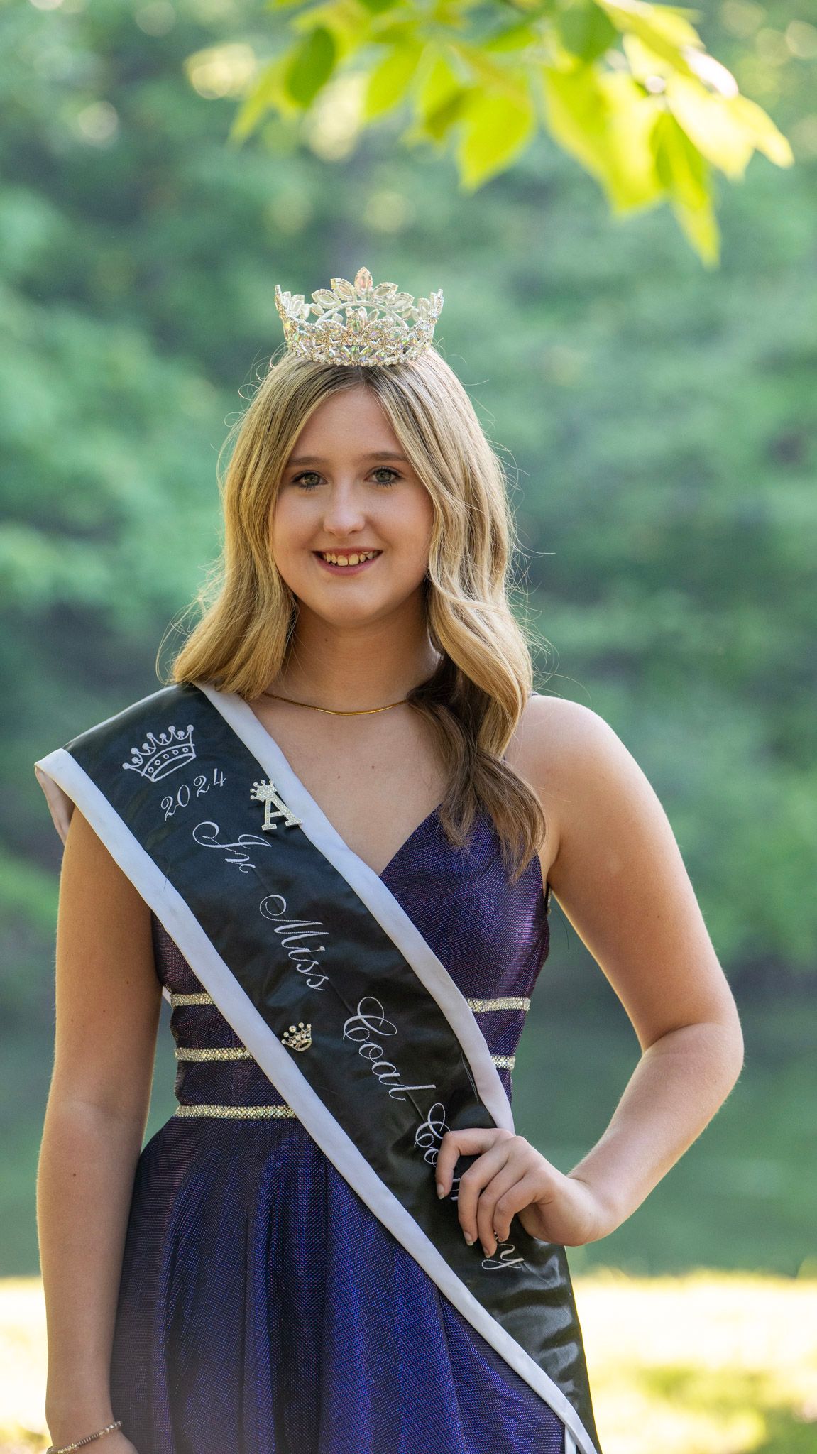 A pageant winner in navy dress poses with crown and sash against a natural backdrop.