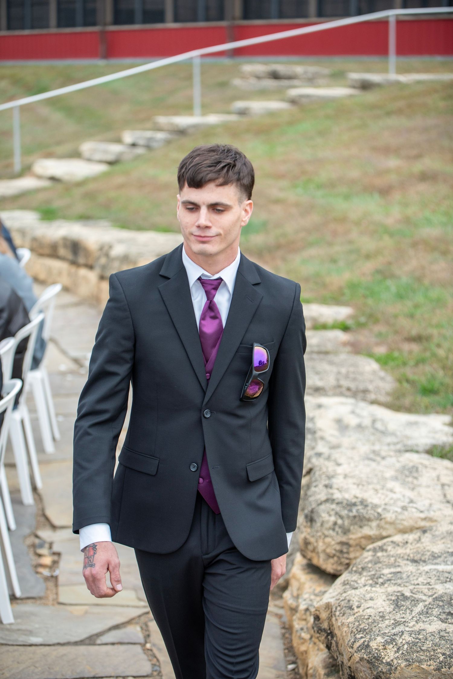 Wedding party member in black suit with purple tie standing on stone steps outdoors.