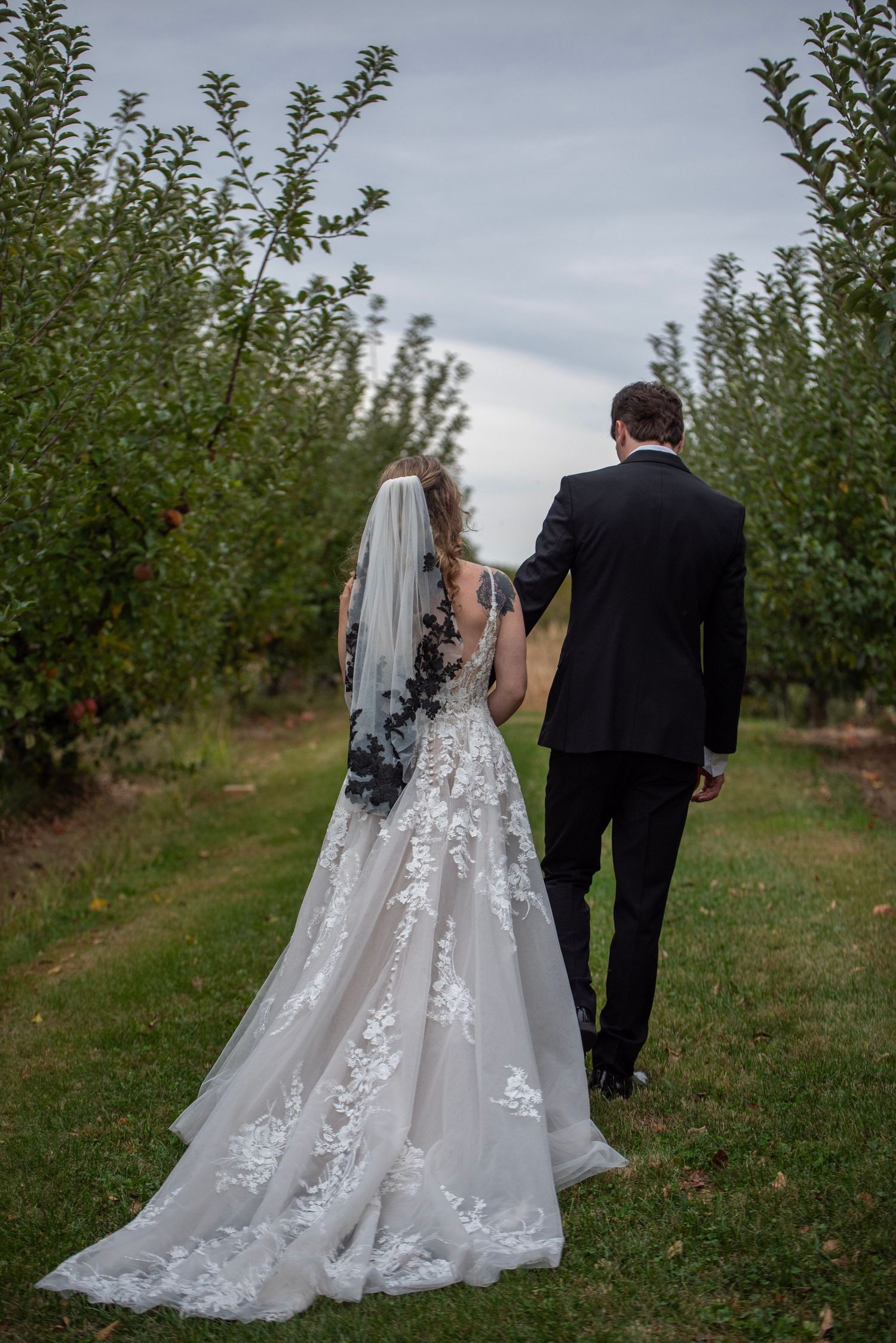 A couple in wedding attire walks through an apple orchard with a flowing lace veil trailing behind.