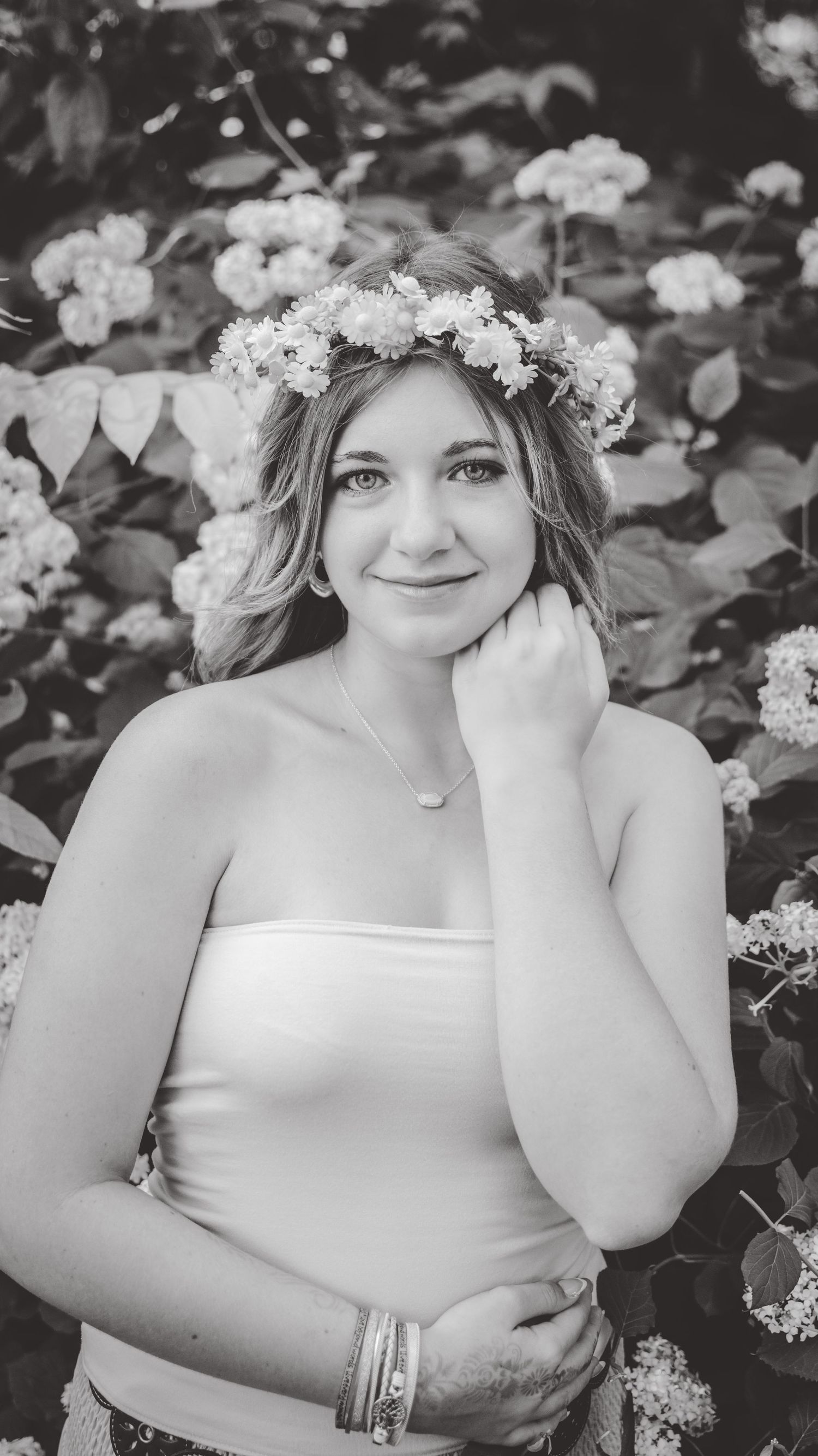 Black and white portrait with white floral crown and strapless dress among flowering bushes.