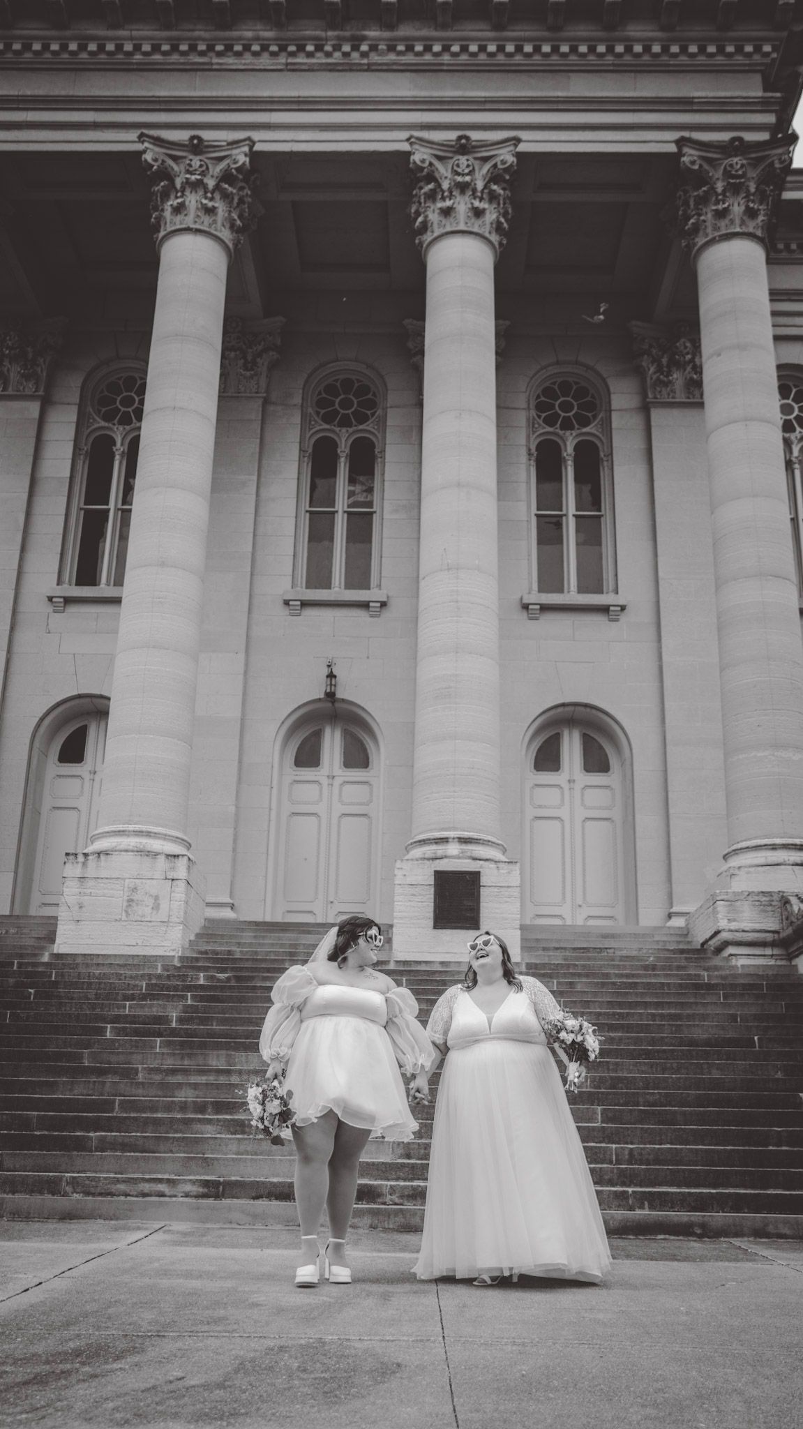 Two people in wedding attire dance and twirl on stone steps in black and white.