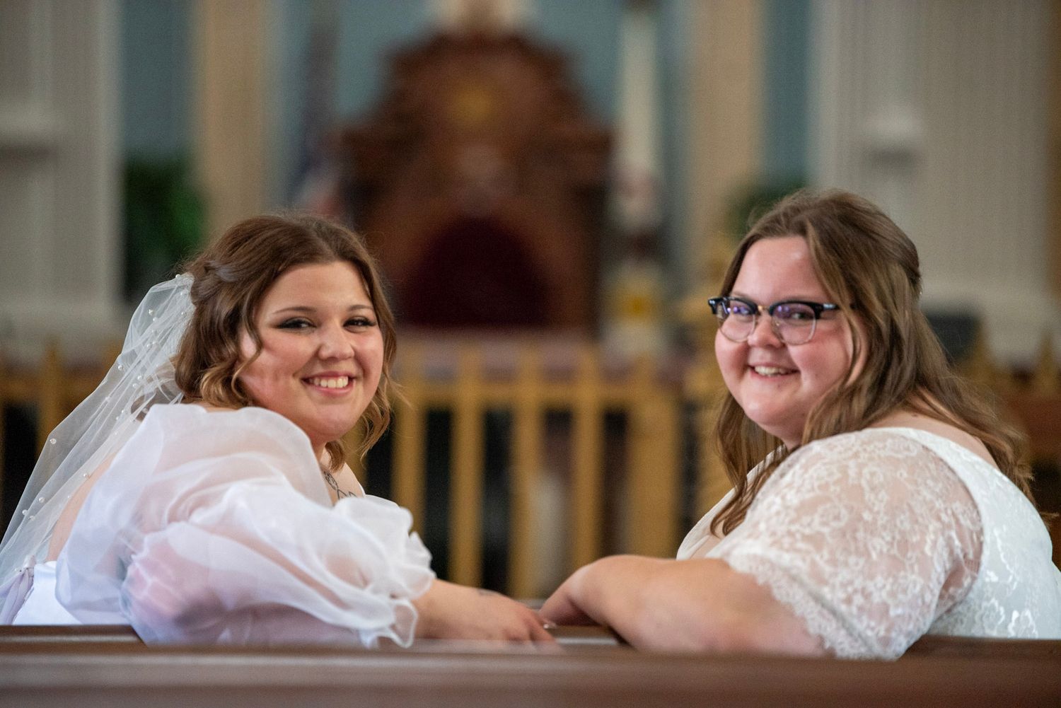 Two brides share a joyful moment while sitting together in a church pew.