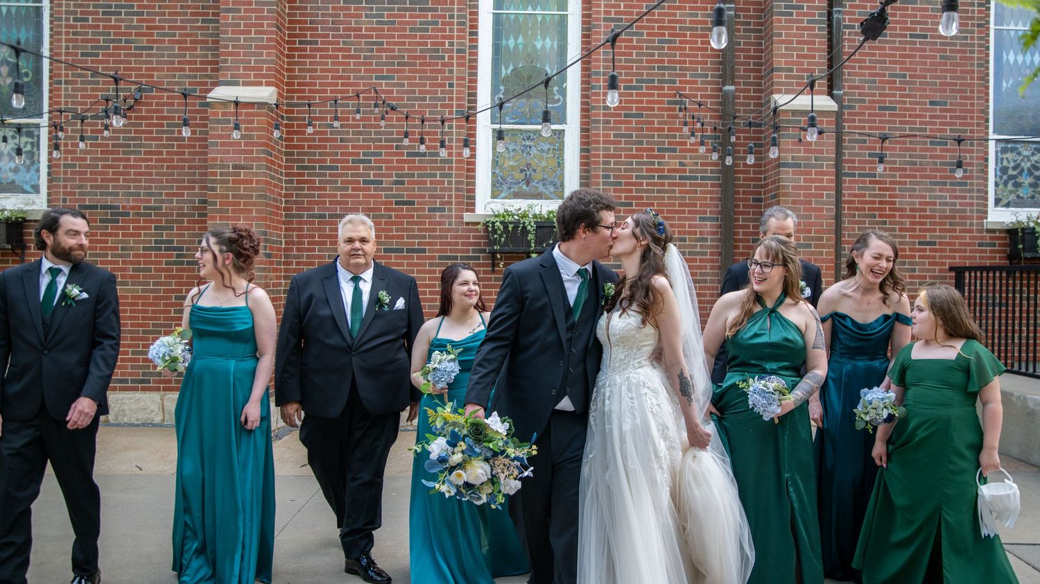 Panoramic wedding party photo showing formal group against brick building backdrop.