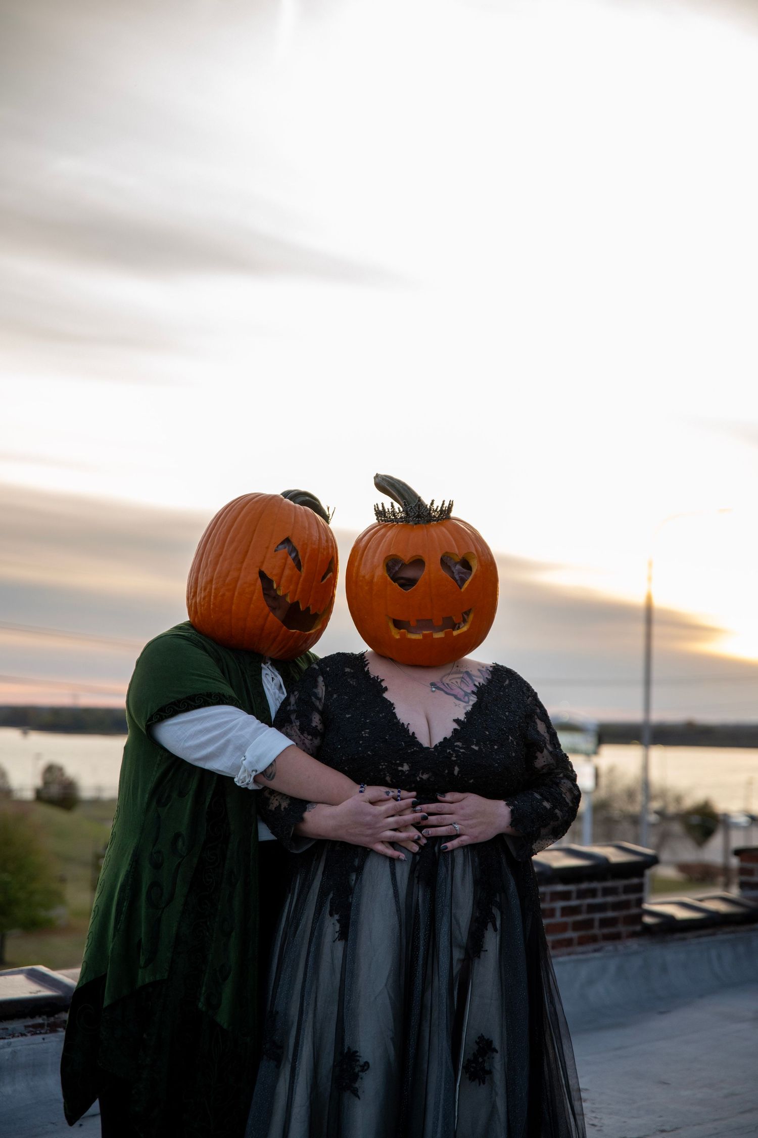 Two figures with pumpkin heads in Victorian clothing embrace on rooftop at sunset.