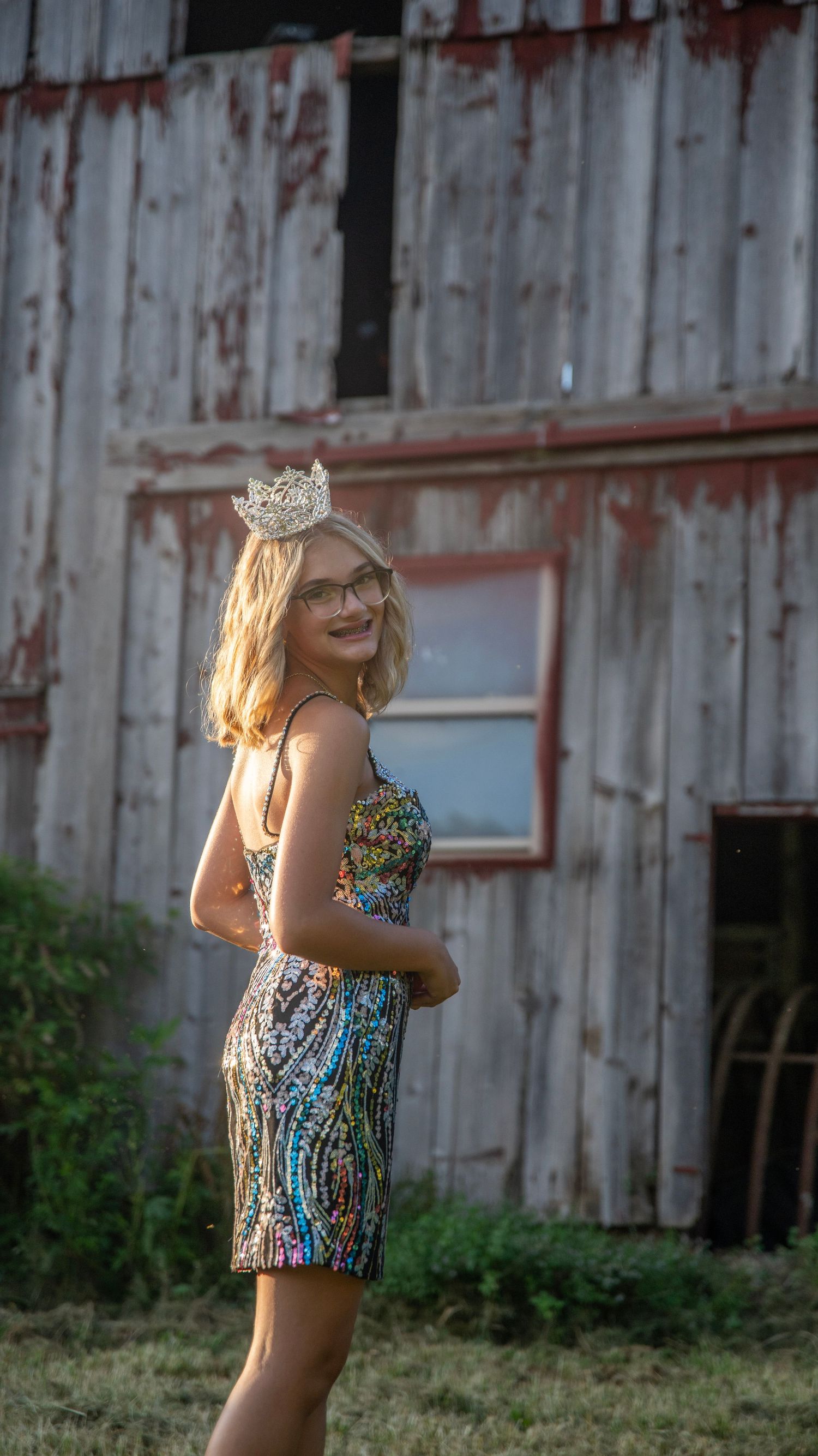 Person in sequined dress and crown poses elegantly against weathered barn siding.
