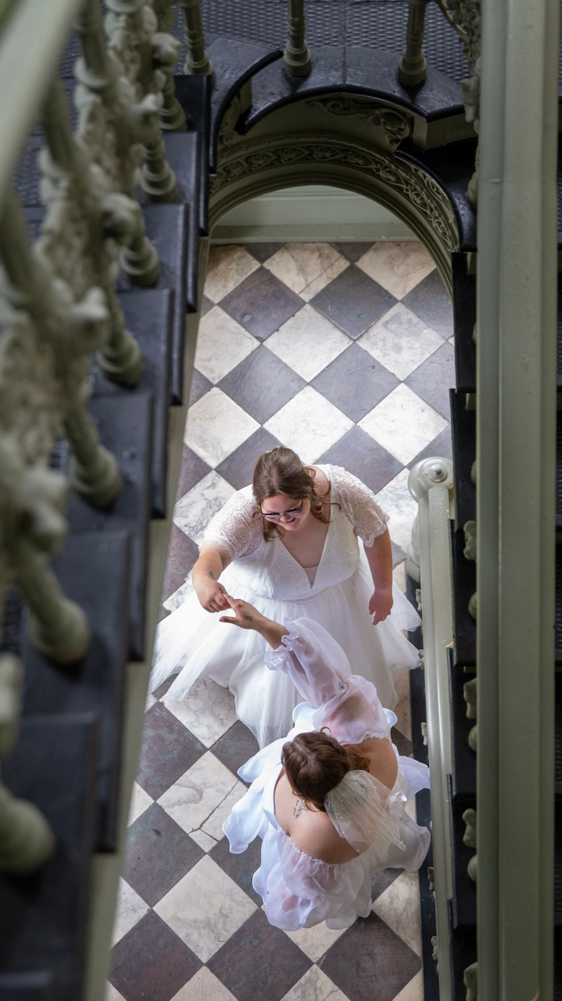 Aerial view of couple in white dresses dancing on a checkered marble floor beneath an ornate archway.