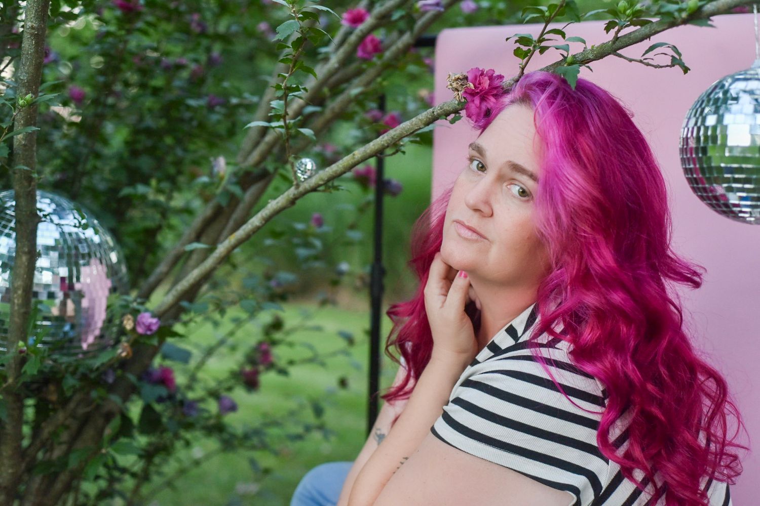 Outdoor photoshoot scene with bright pink hair and striped clothing against natural greenery.