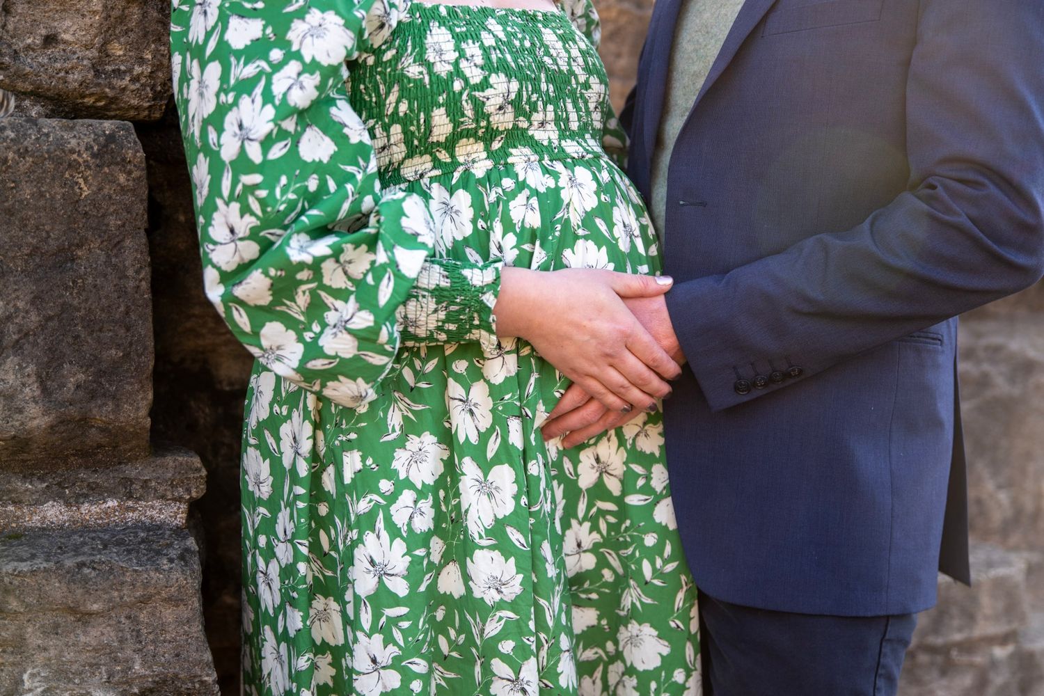 Romantic couple embracing outdoors, with woman in green floral dress against stone wall.