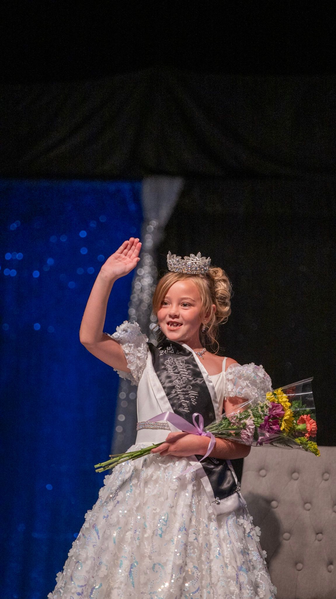 A young pageant winner in a white dress and crown waves to the audience on stage.