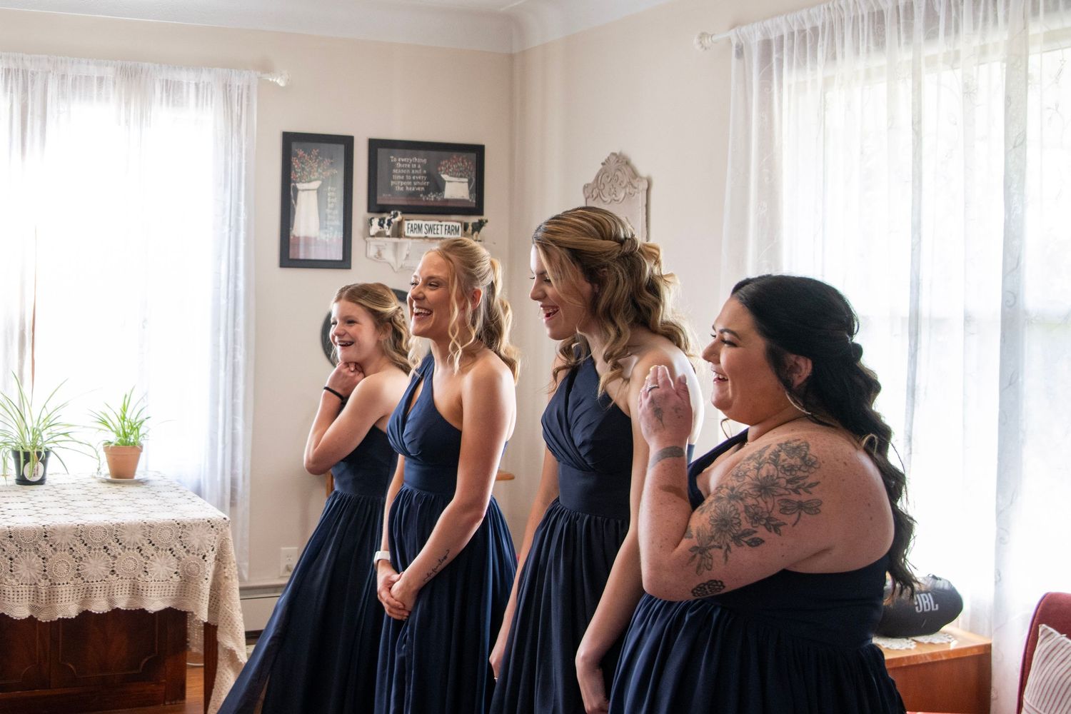 Bridesmaids in navy blue dresses gather excitedly in a sunlit room before the wedding ceremony.