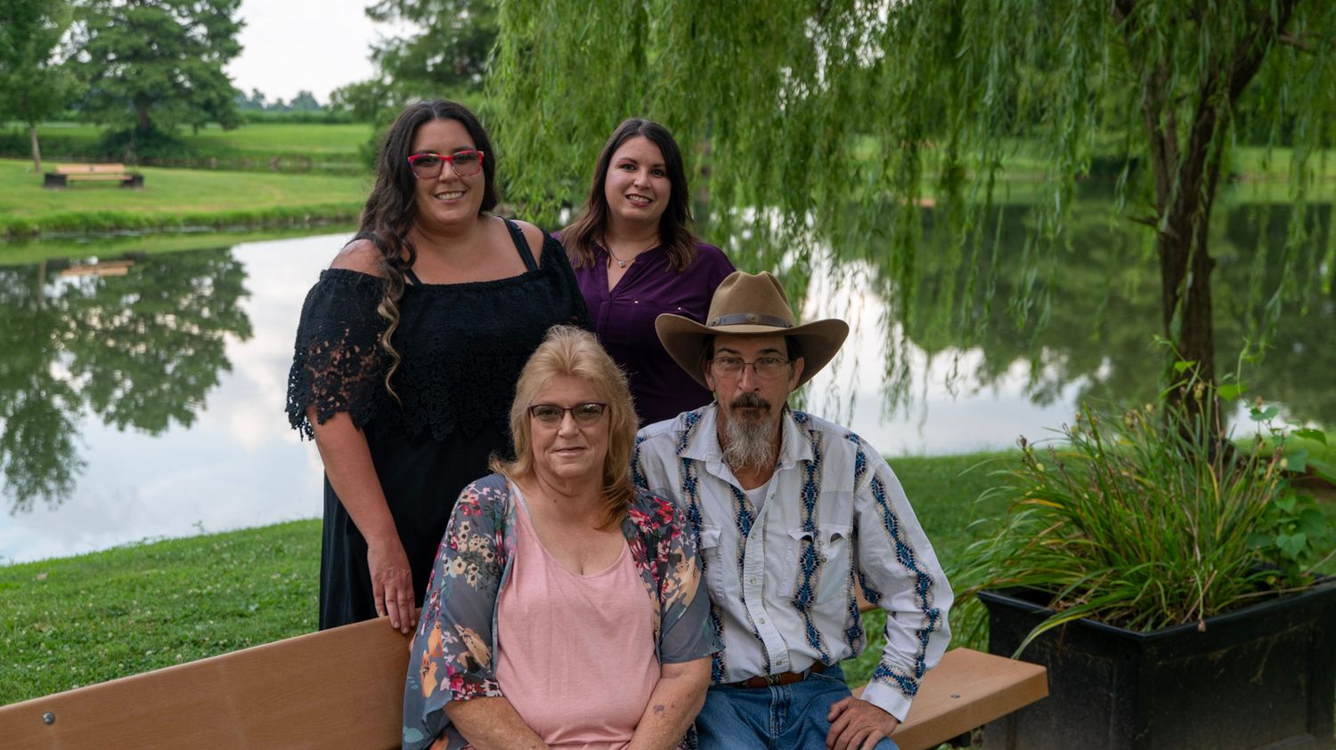 People gathered on wooden benches under a weeping willow tree near a pond.