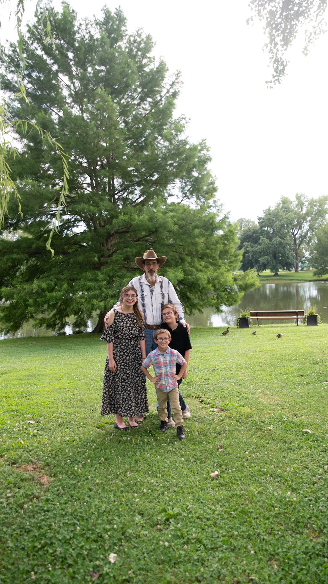 A group stands together on a grassy lawn near a lake with trees in the background.