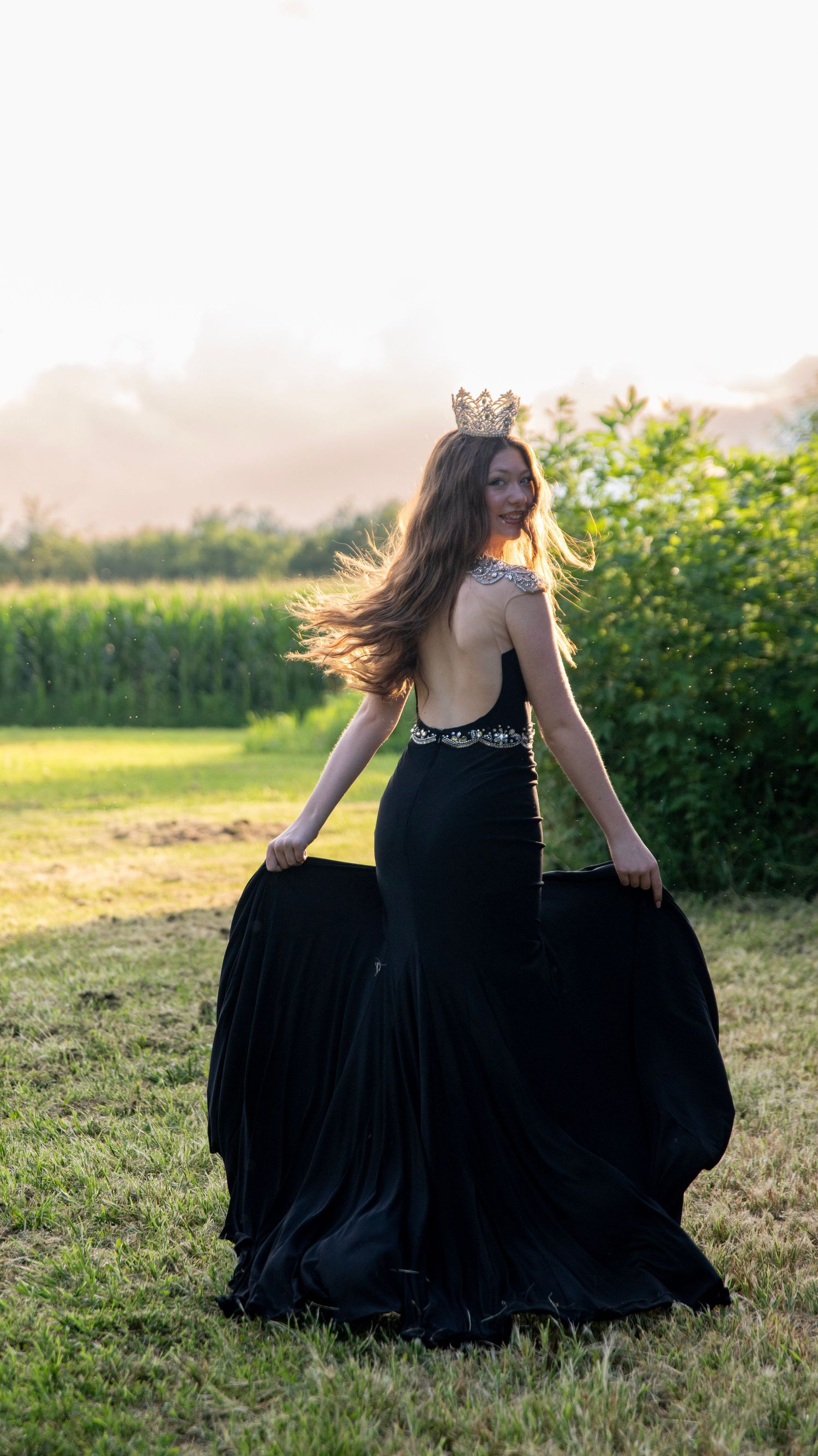 A black dress twirls gracefully in a sunlit field with greenery in the background.