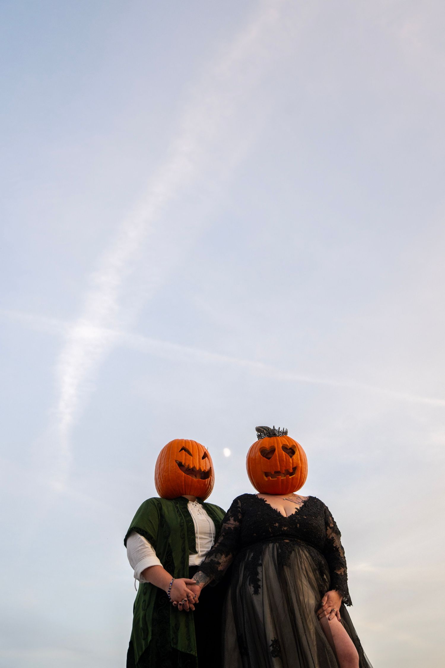 Two figures in Victorian clothing with pumpkin heads pose against bright sky.