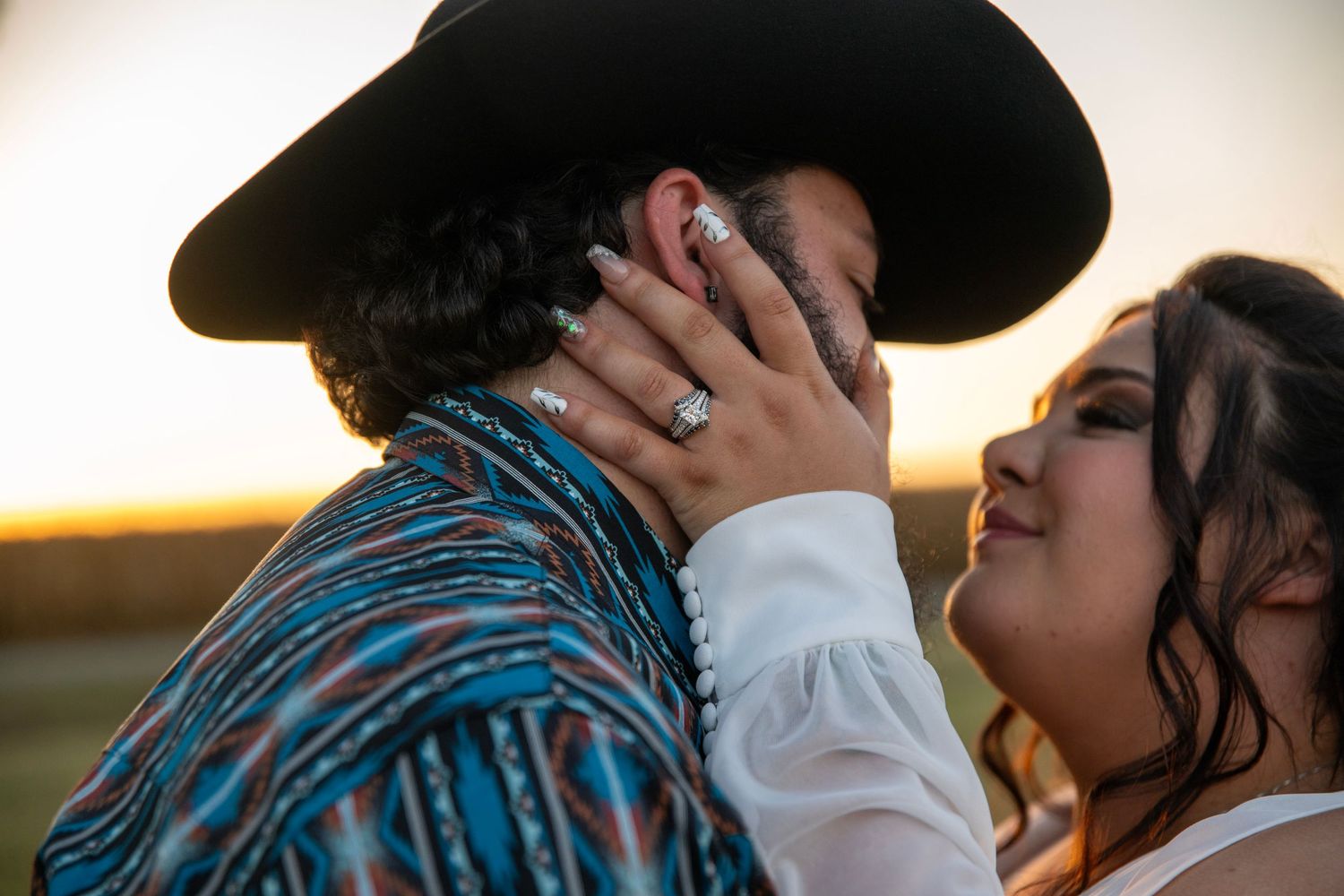 Romantic sunset portrait of a couple with hands caressing face, one wearing a western hat.