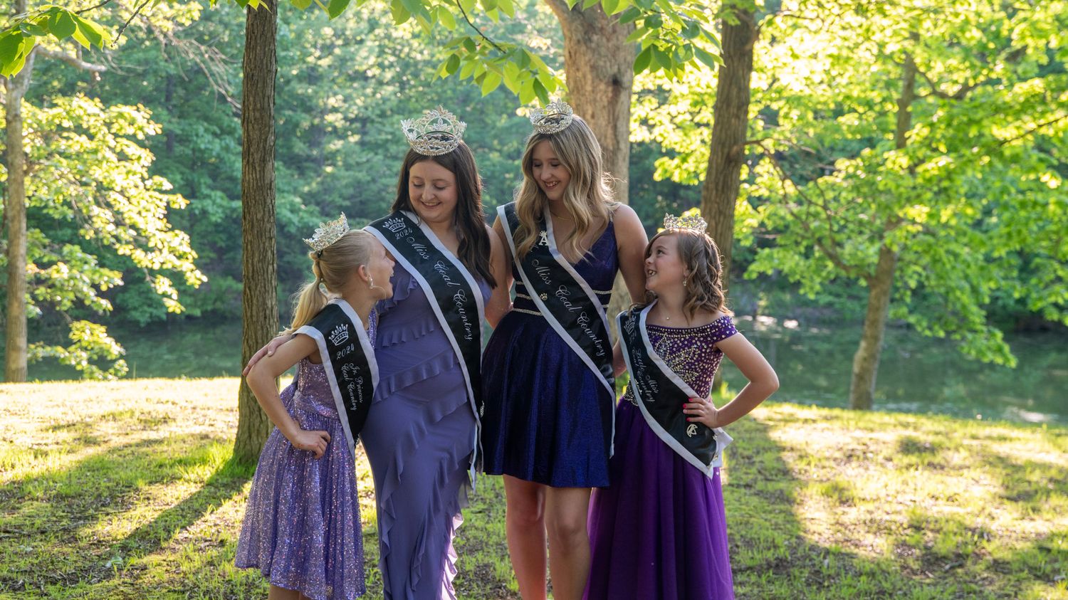 Beauty pageant participants in matching purple gowns stand together among trees.
