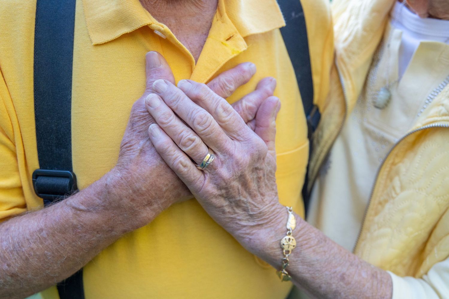 Elderly hands clasped together against a bright yellow shirt with black suspenders visible.