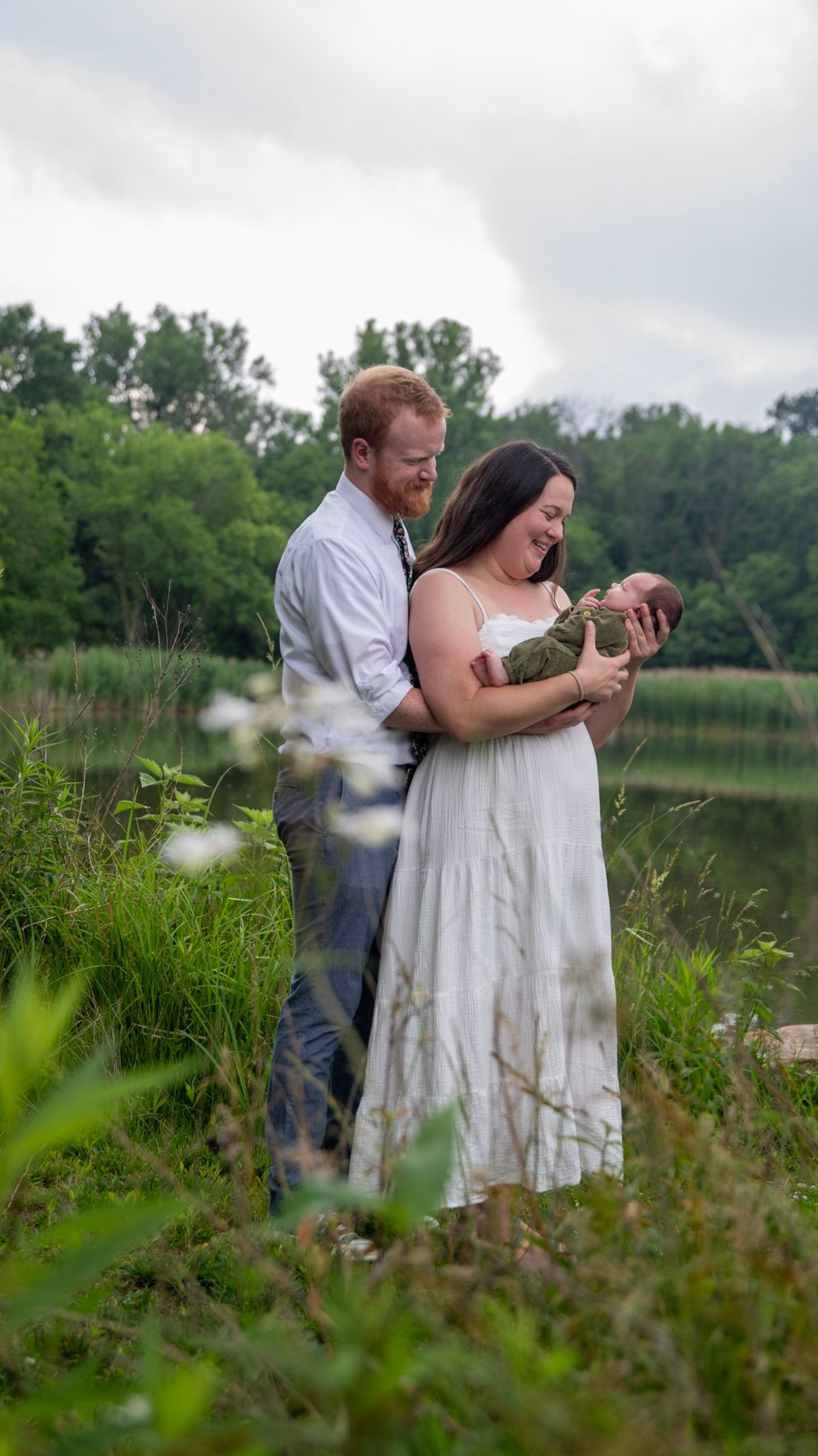 A couple stands together in tall grass near a peaceful lake on an overcast day.