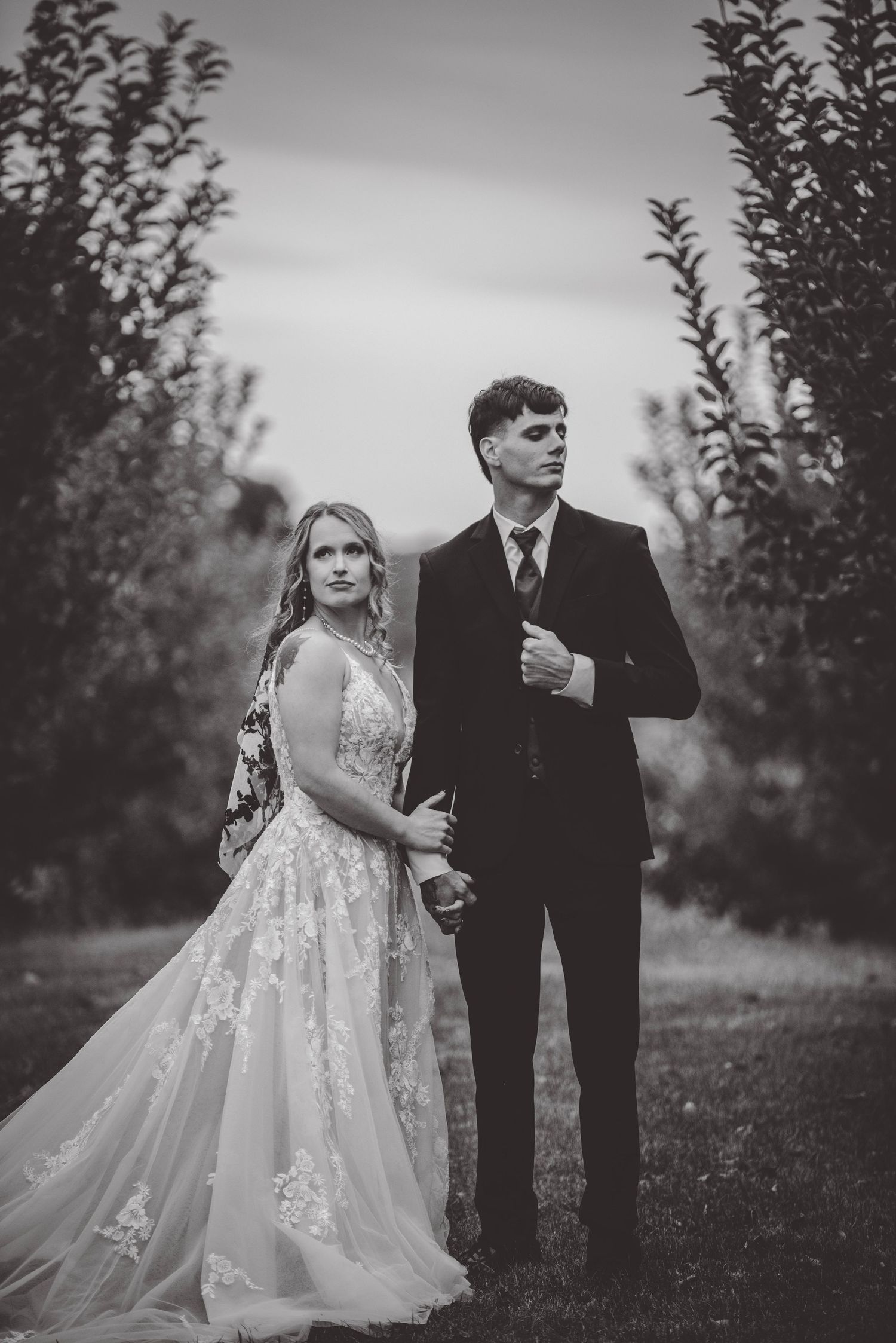Black and white portrait of a couple in wedding attire standing among apple trees at dusk.