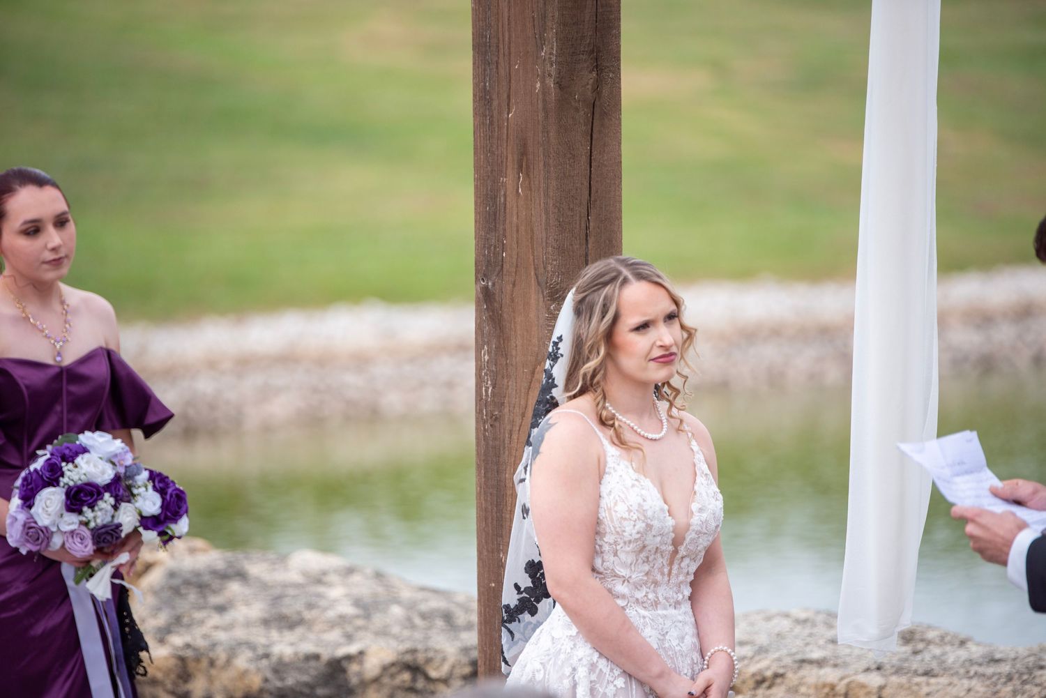 Wedding ceremony participants standing under decorated pergola by the water.