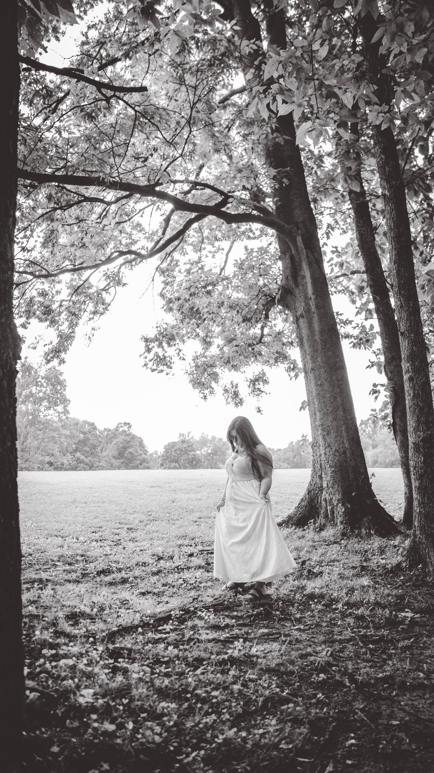 Black and white photo of a person in white dress standing beneath tall trees.