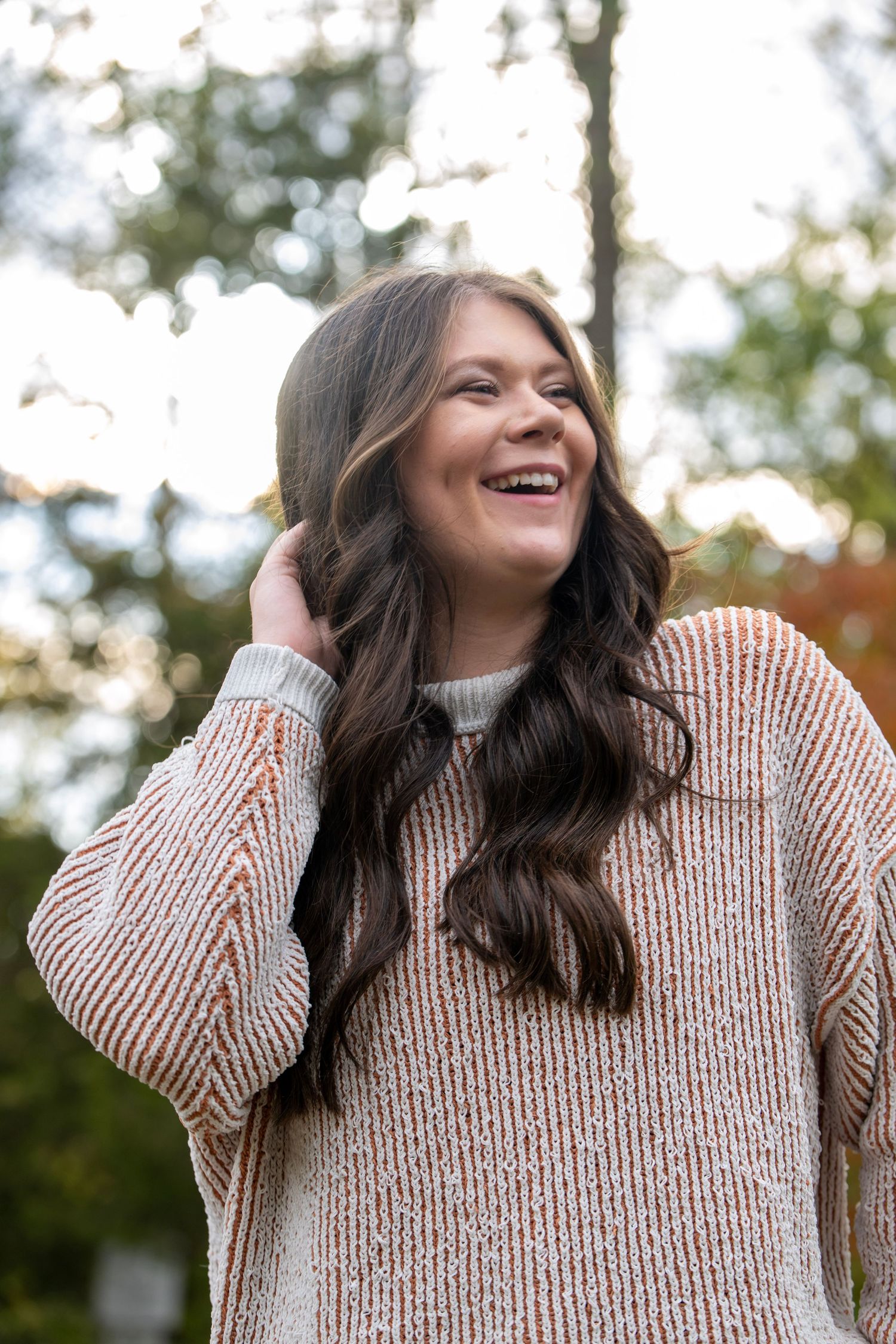 Person wearing beige knit sweater laughing joyfully outdoors with autumn trees in background.