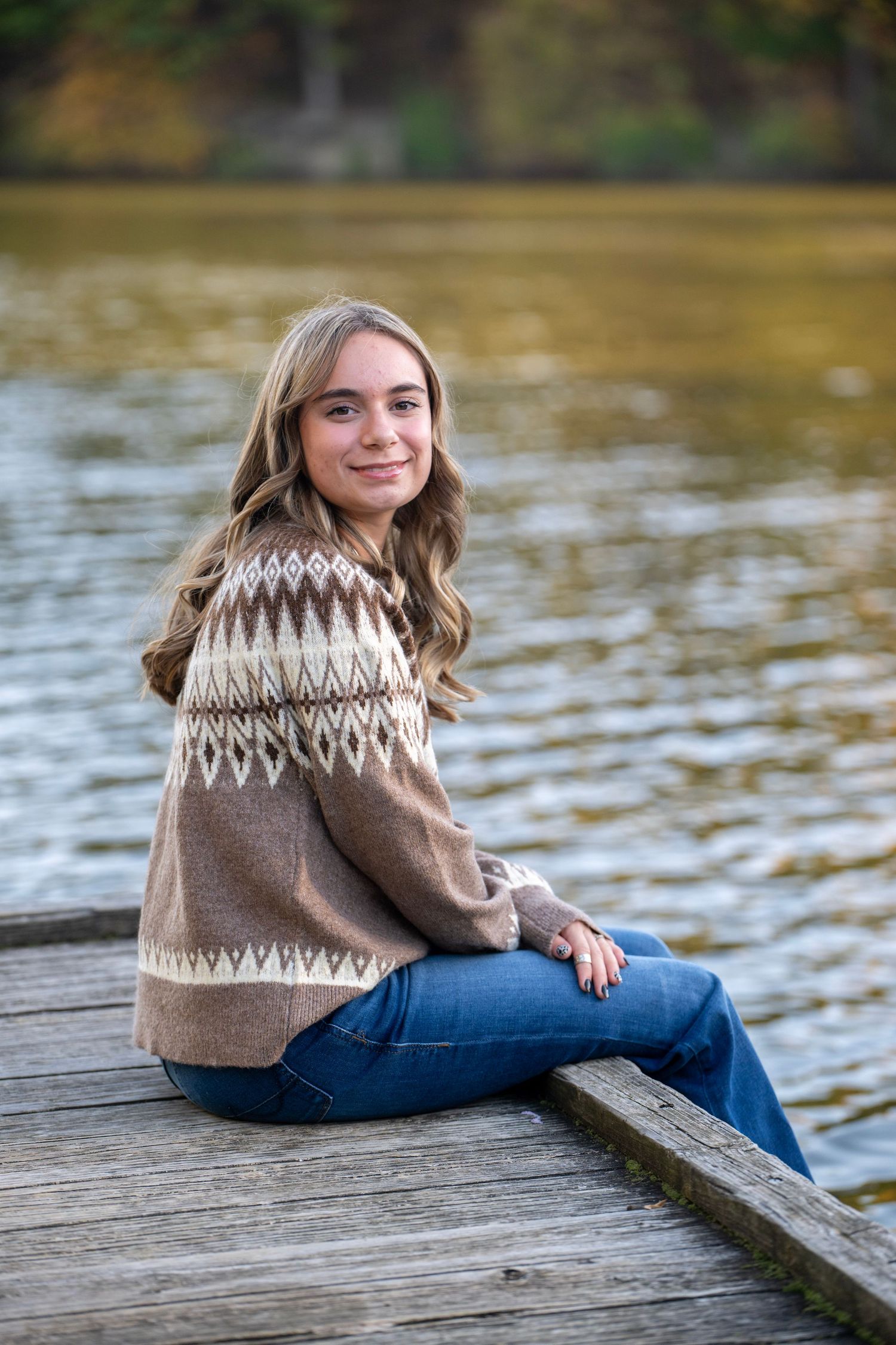Person in brown patterned sweater sitting on wooden dock by water.