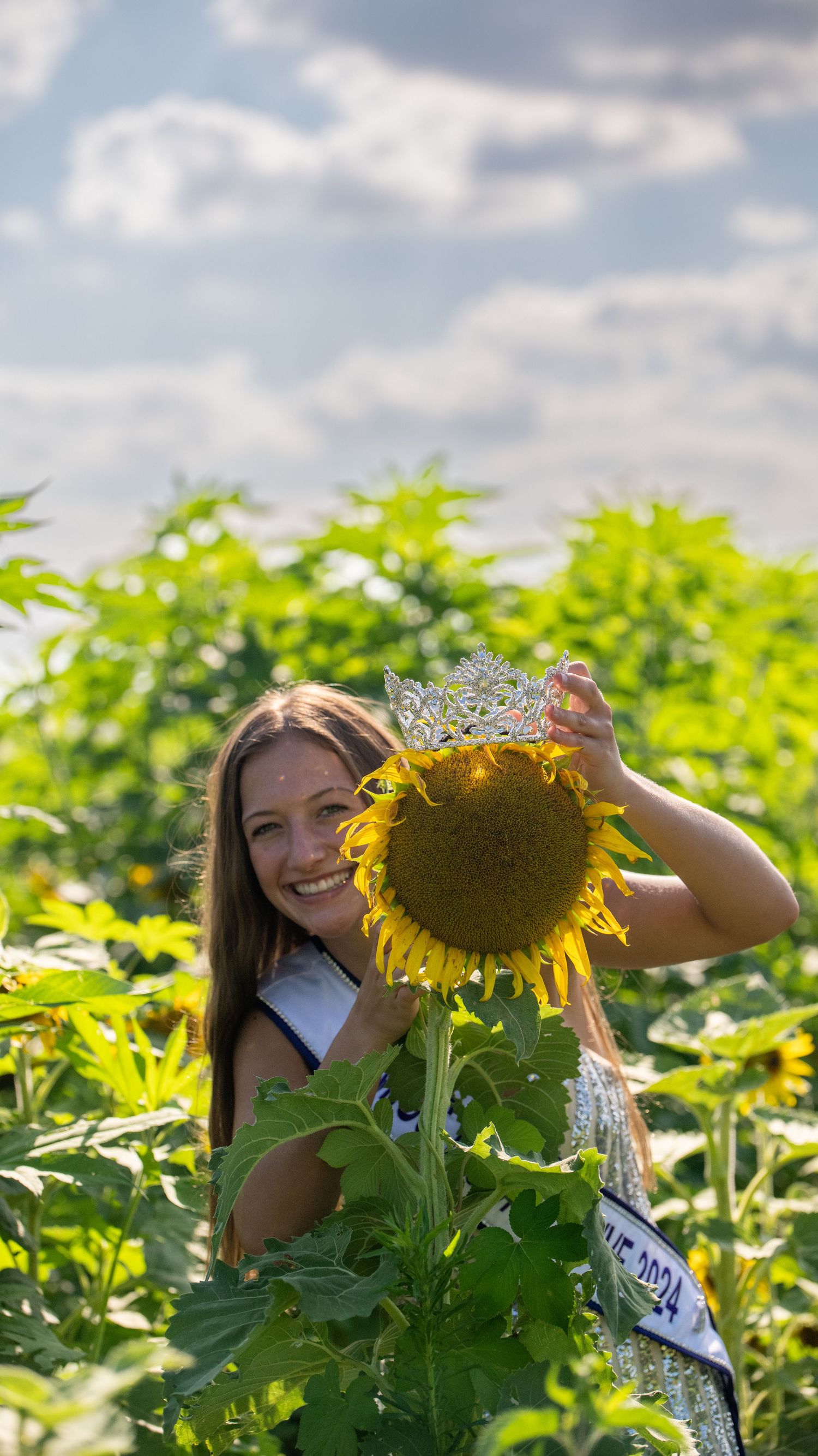 A pageant winner places her crown on a large sunflower in a sunny field.