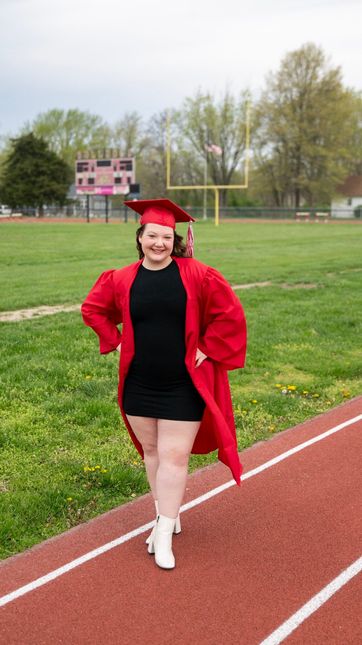 Graduate in red cap and gown poses playfully on a running track with football field in background.