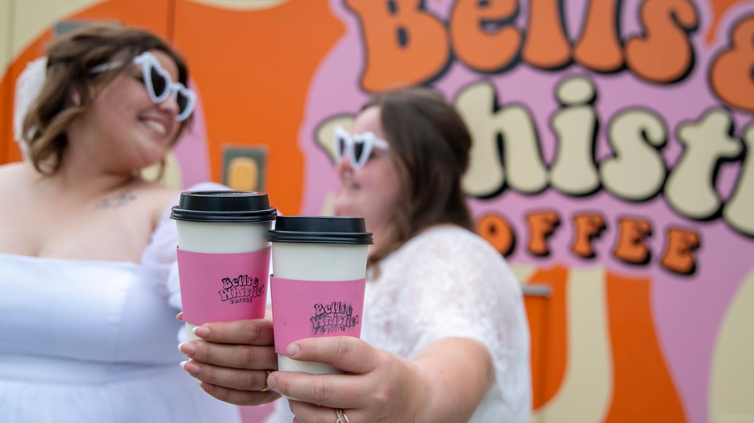 People holding pink coffee cups in front of colorful Dunkin Donuts signage.