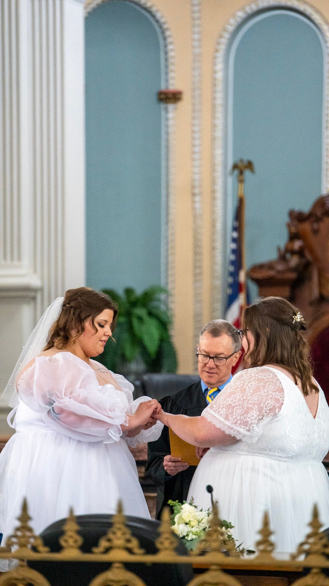 Two people in white dresses signing documents at a courthouse ceremony.