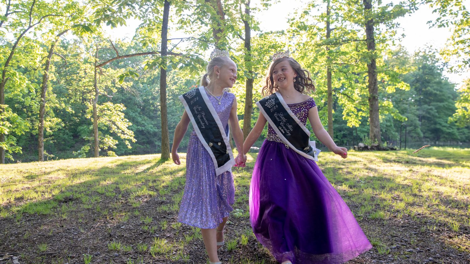 Pageant contestants in purple formal wear interact playfully in a wooded outdoor setting.