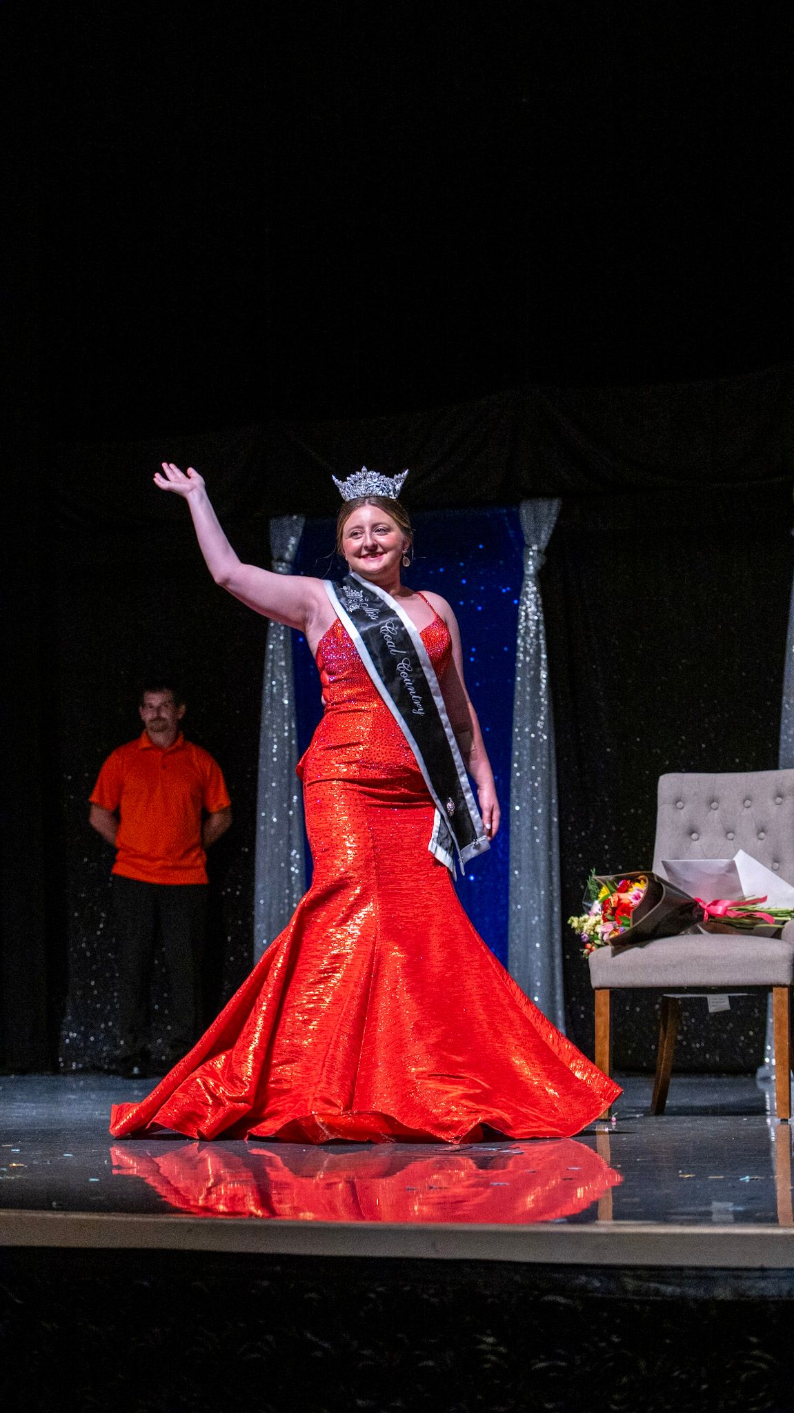A performer in a dramatic red ball gown twirls on stage during a pageant show.
