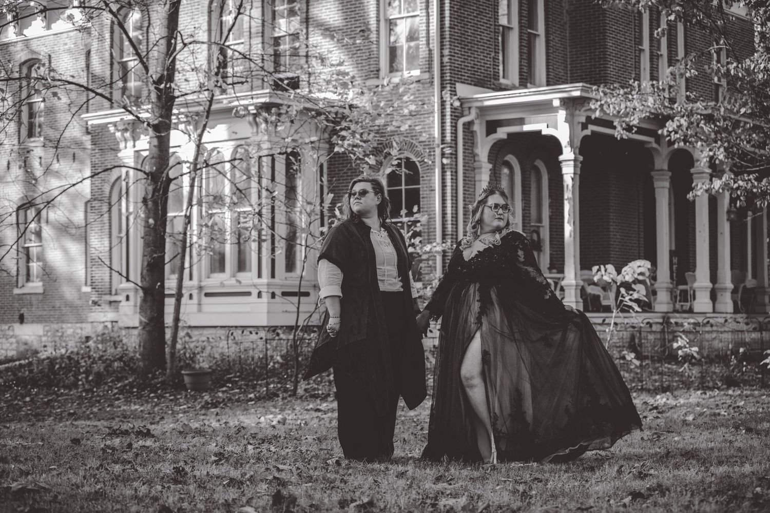 Black and white image showing figures in period dress near a Victorian mansion.