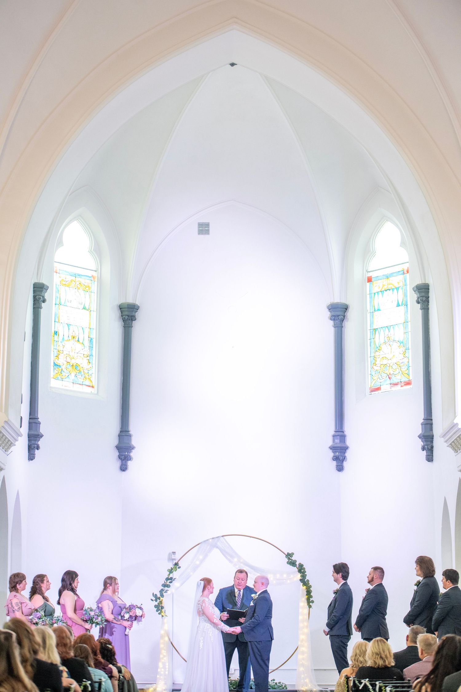 A wedding ceremony takes place under soaring cathedral arches with stained glass windows and a circular floral backdrop.