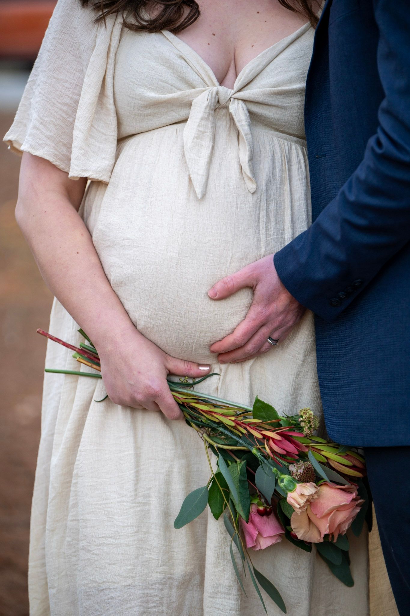 A pregnant person in a cream dress holds pink roses while a hand gently rests on their belly.