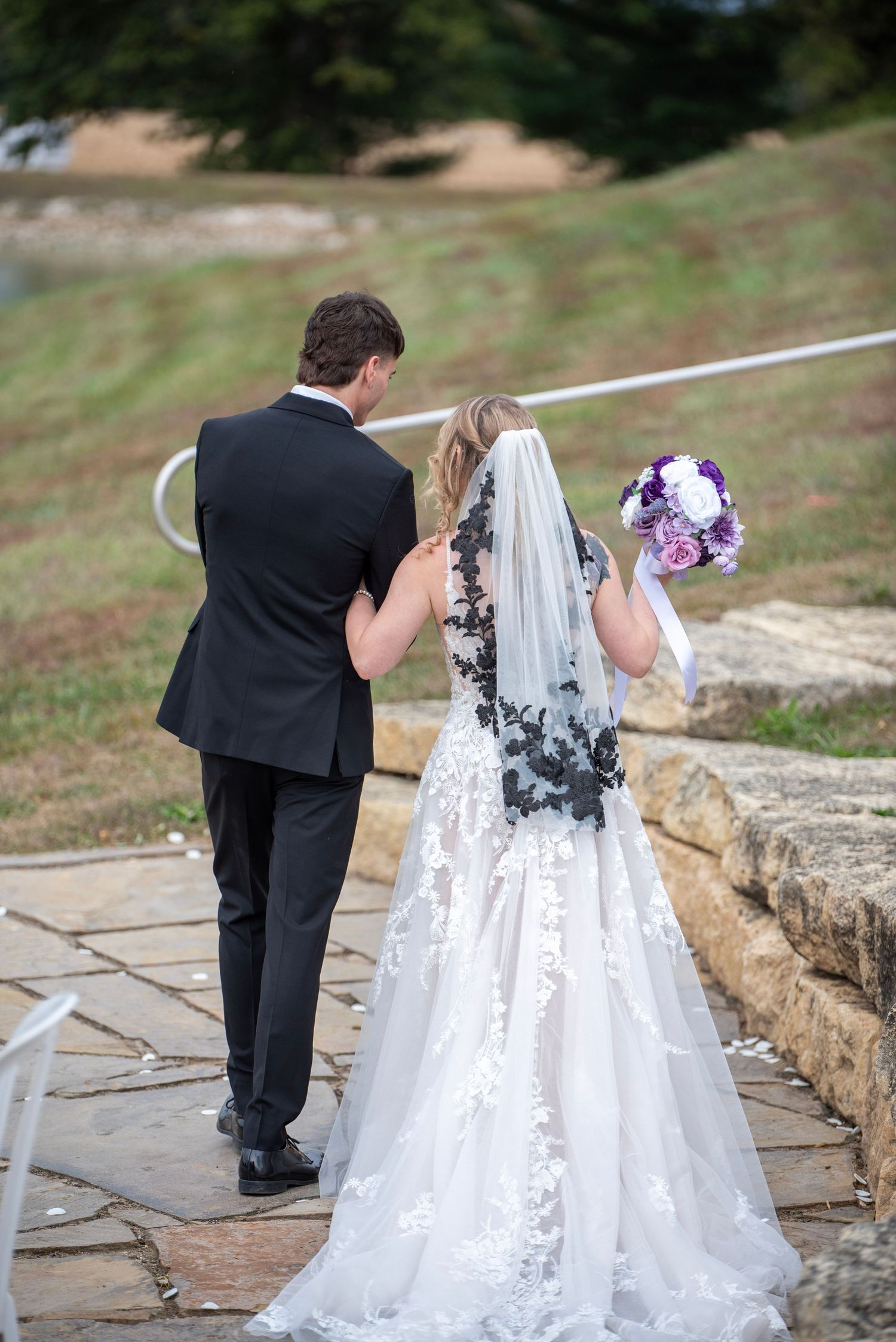 Couple walking along stone pathway with purple floral bouquet at outdoor wedding venue.