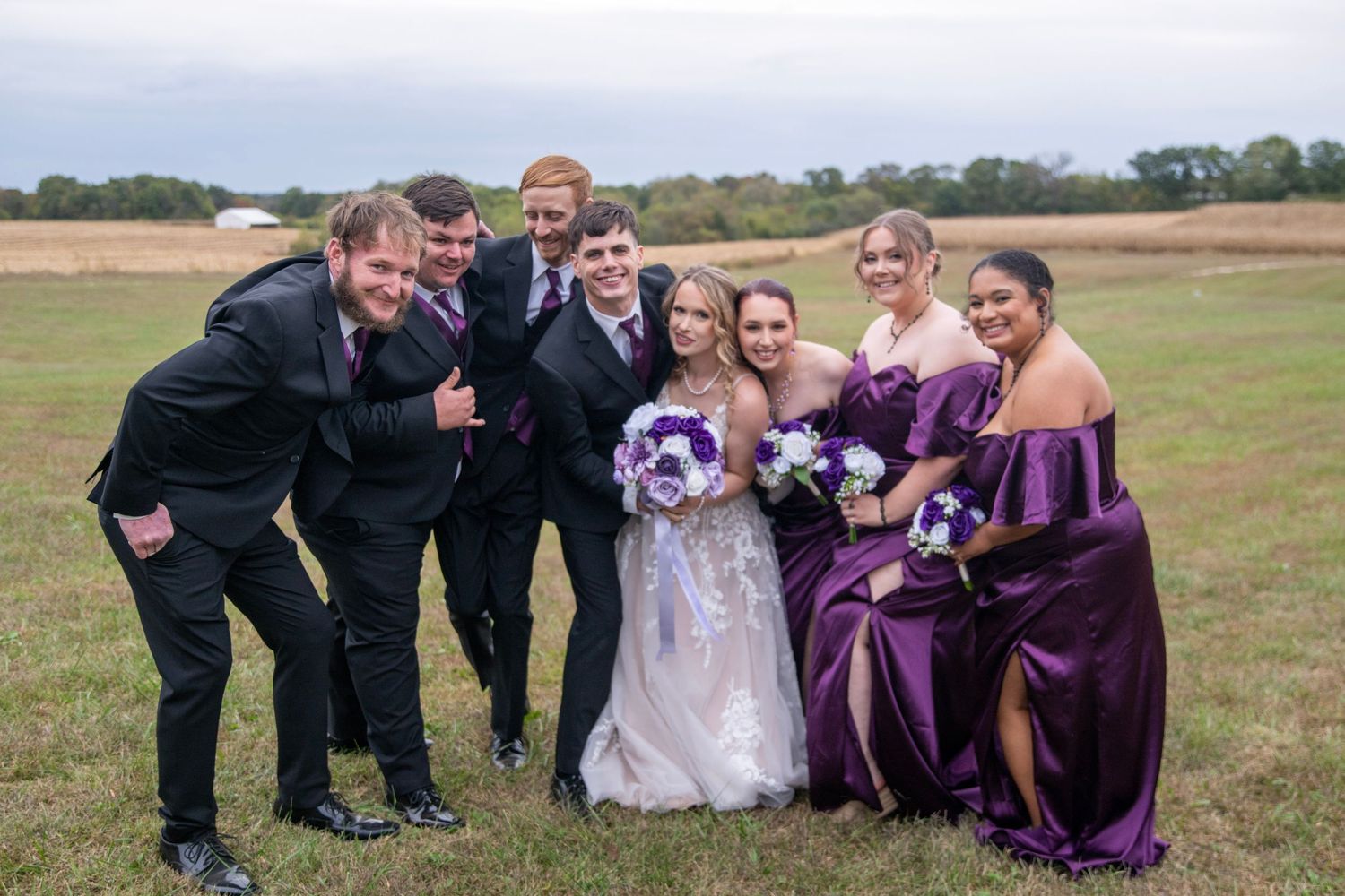 Wedding party shares playful moment outdoors with purple and white floral bouquets.