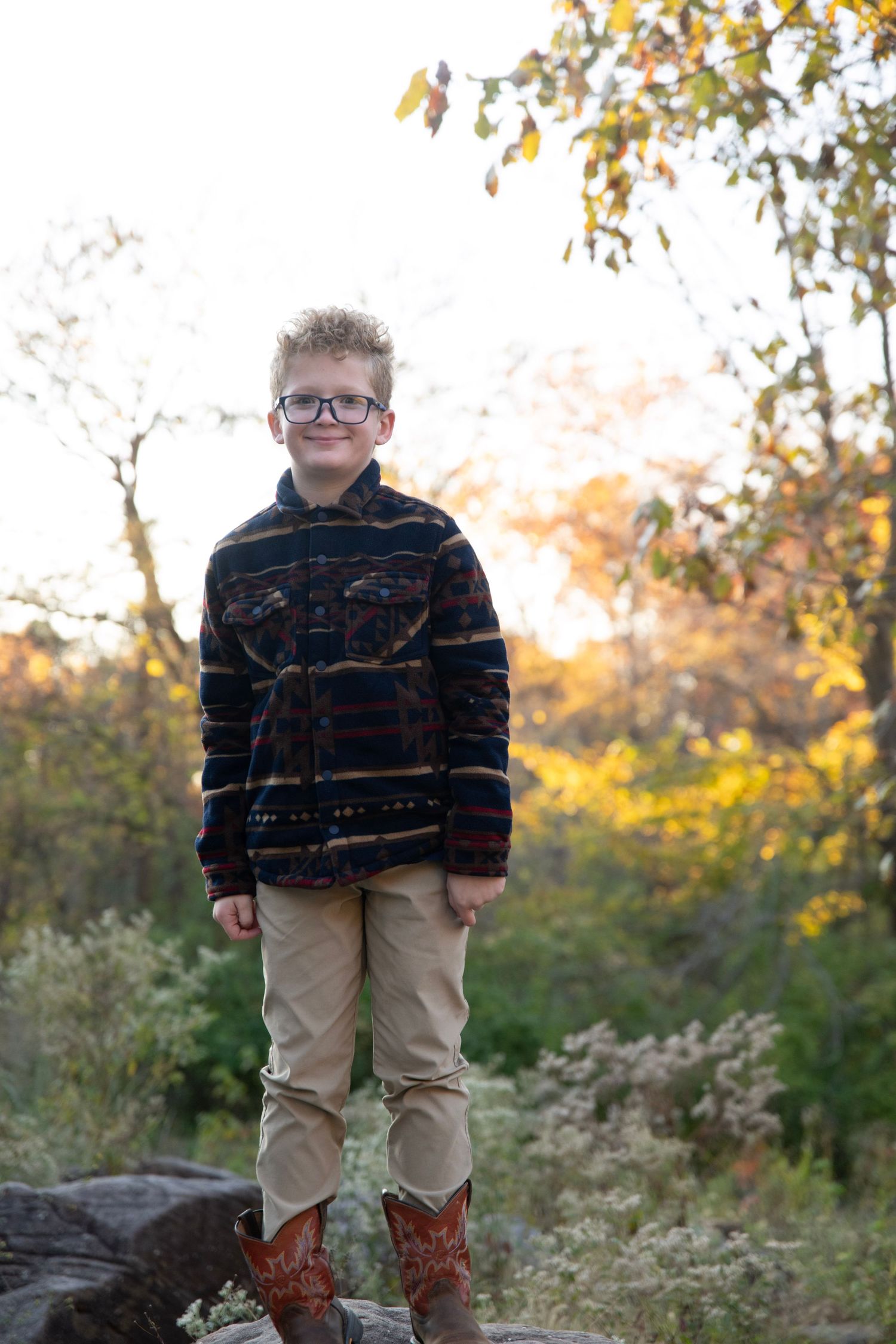 A child stands on rocks wearing khaki pants and a dark sweater against an autumn backdrop.
