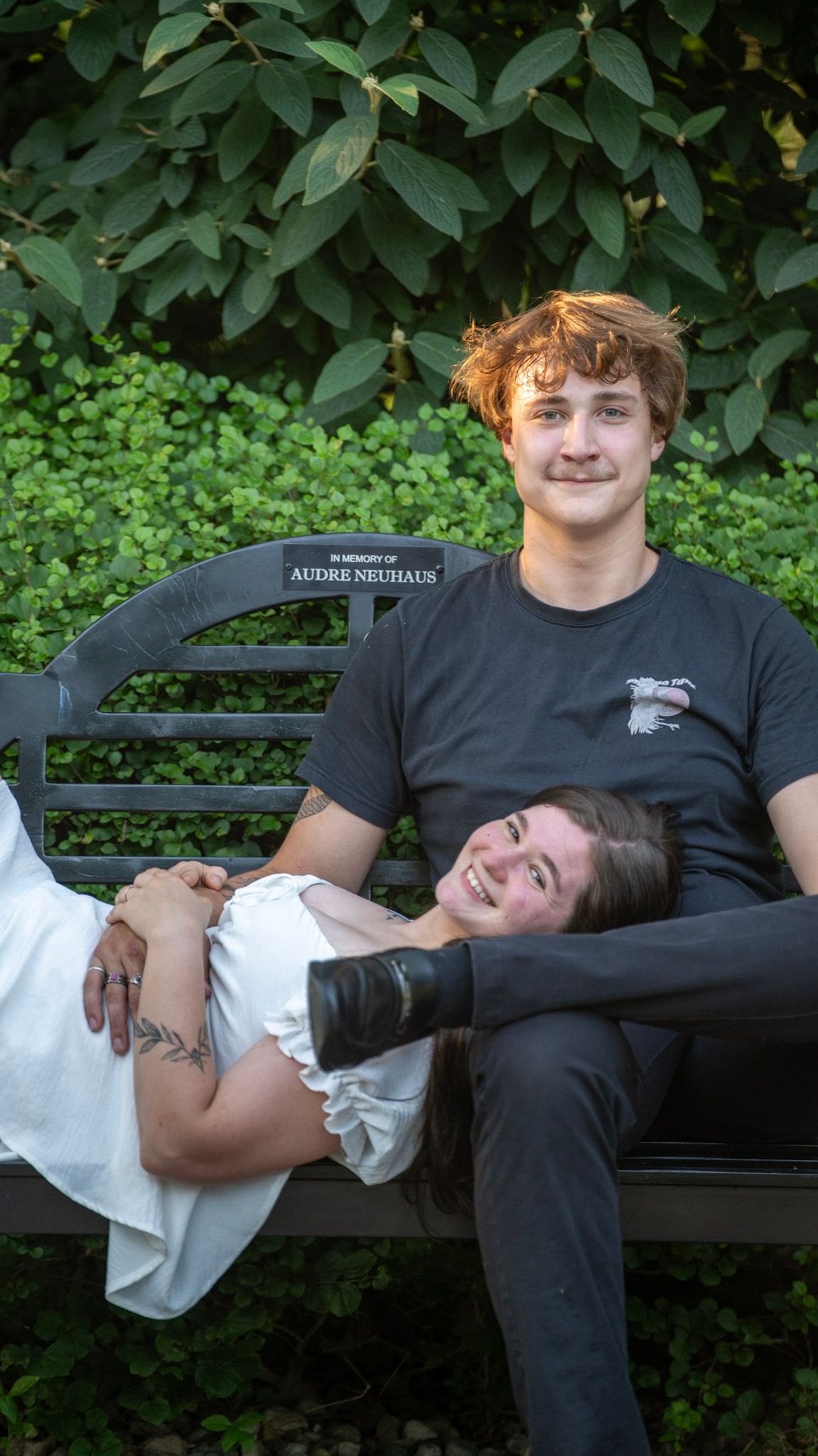 A couple lounges together on an outdoor bench against a leafy backdrop.