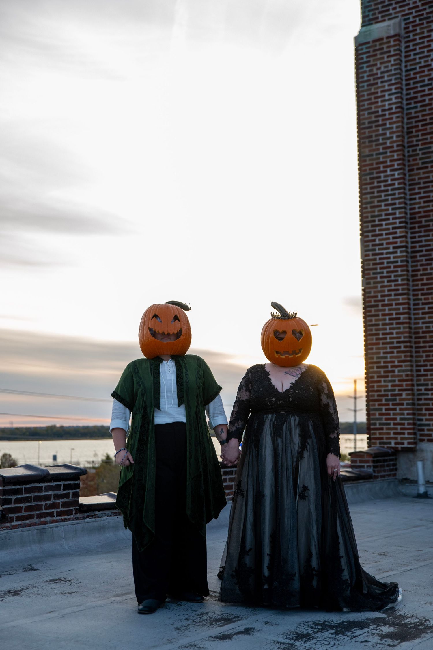 Two figures in Victorian dress wearing pumpkin heads pose against sunset sky on rooftop.
