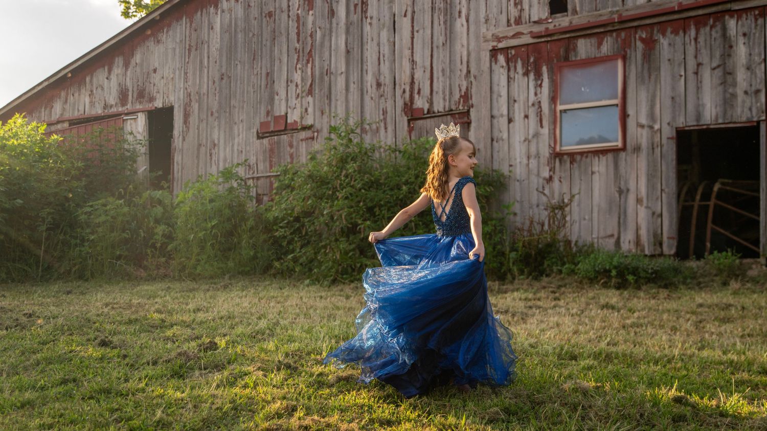 Person in flowing blue gown twirls in front of weathered barn at sunset.