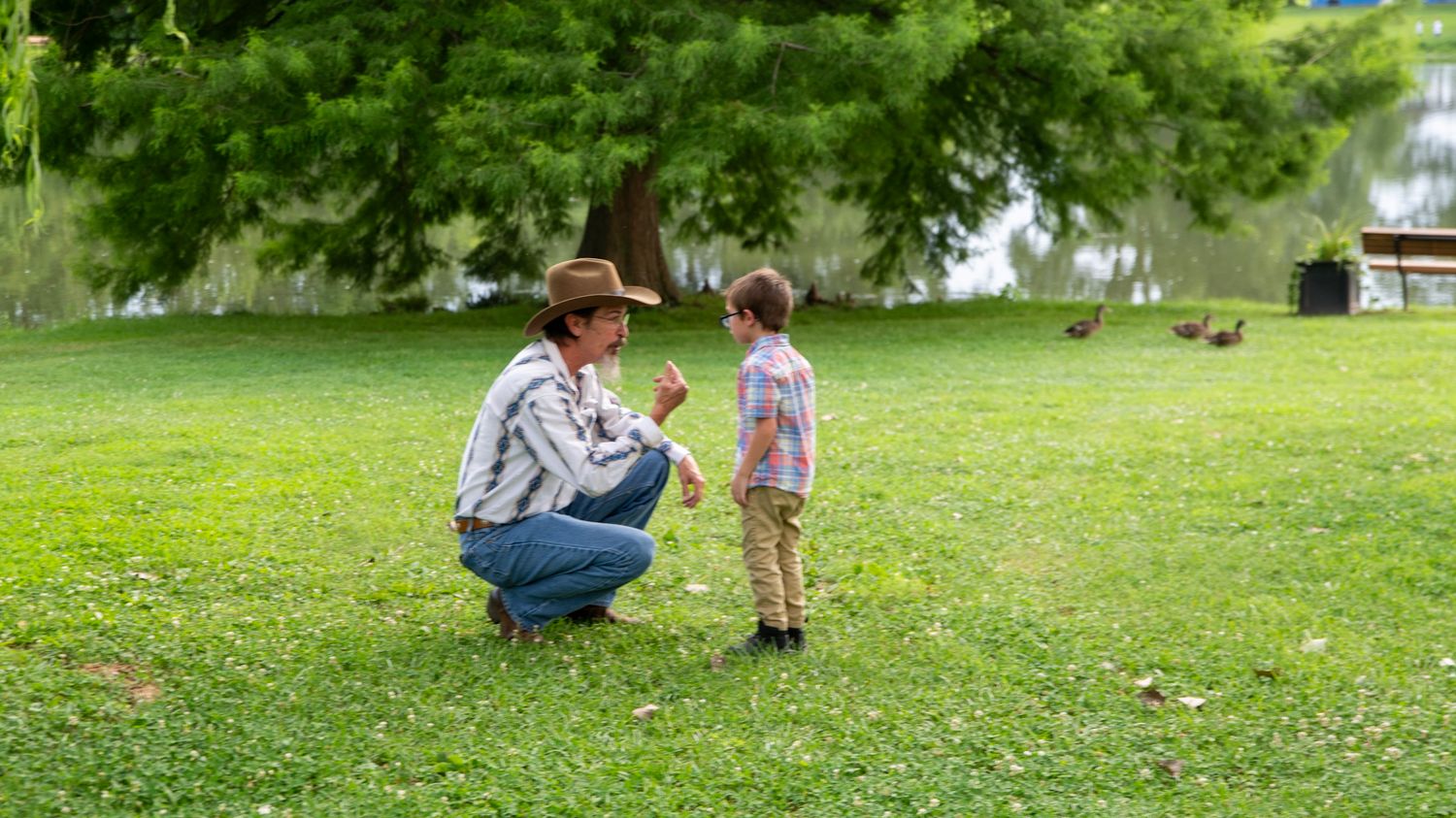 Two figures interact on grassy lawn with pond and trees in background.