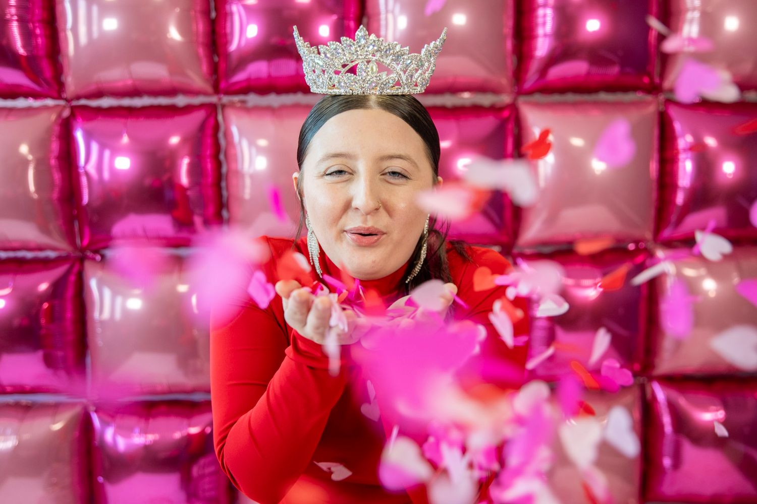 Pink confetti shower over red dress and crown against metallic backdrop.