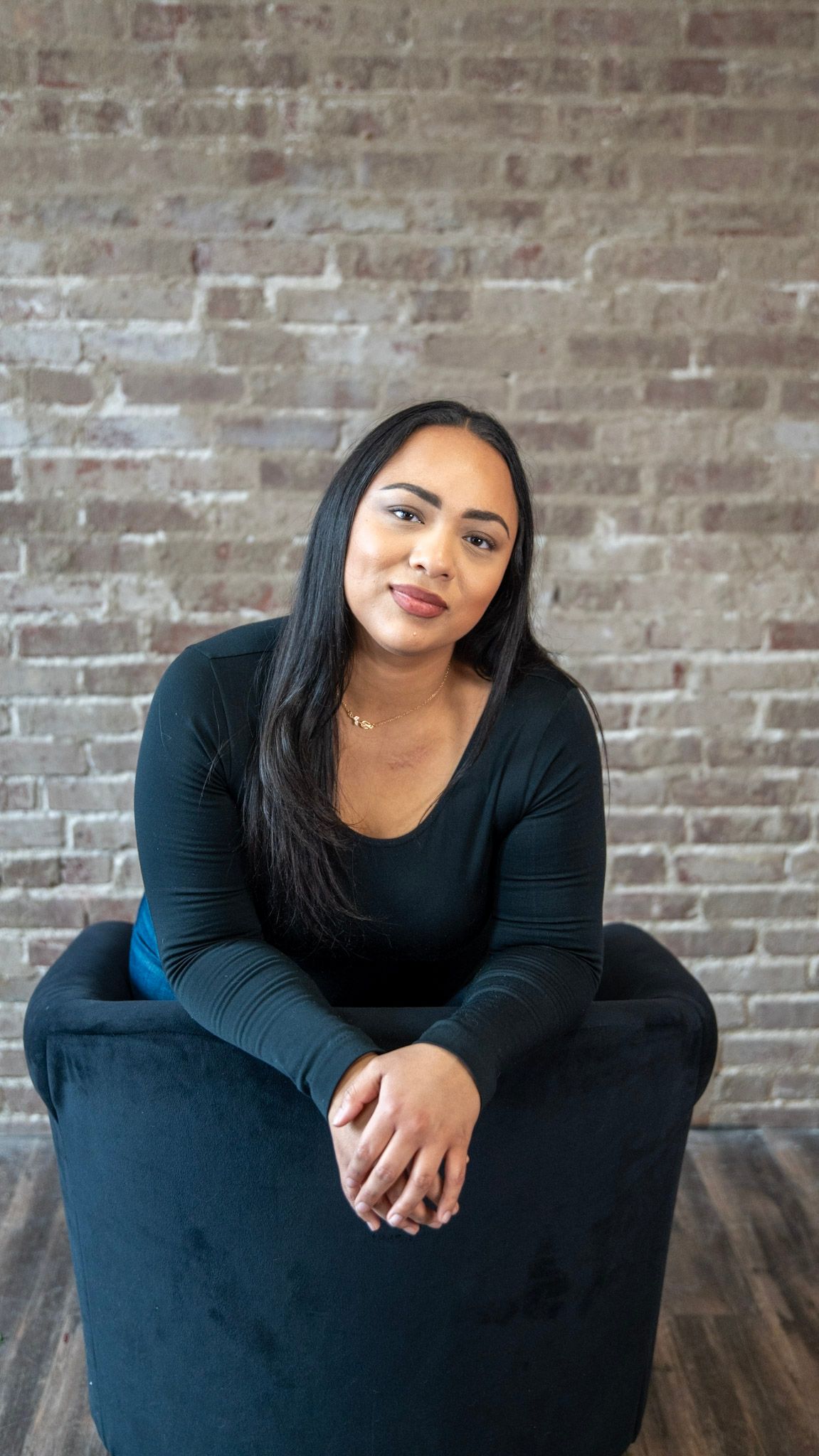 Woman in black top sitting casually against exposed brick wall interior.