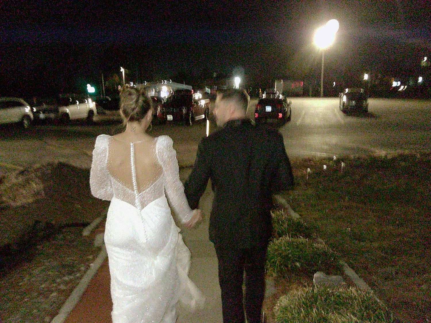 A couple walks hand in hand at night in wedding attire along an illuminated path.