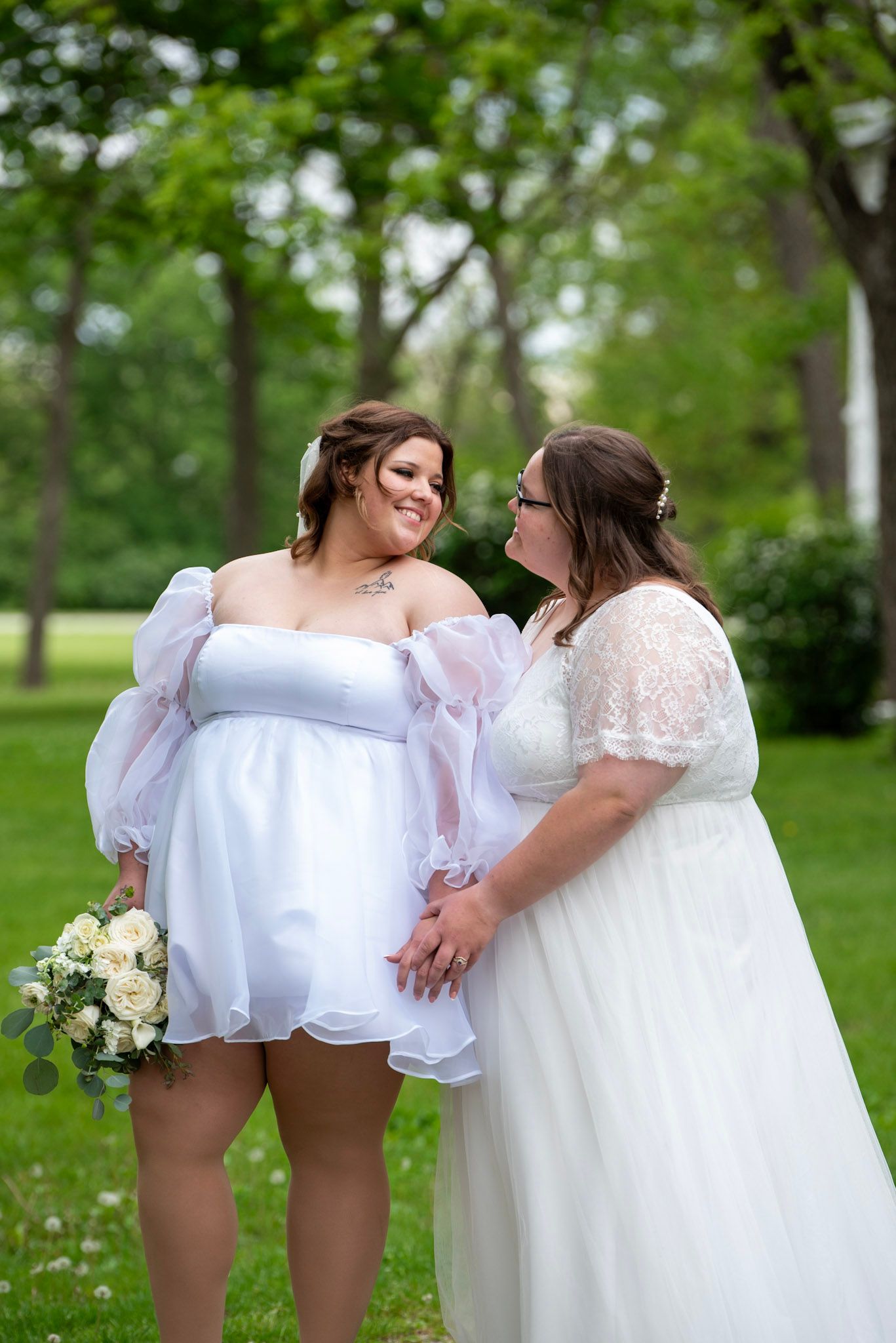 Two people in white dresses share a playful moment in a lush garden setting.